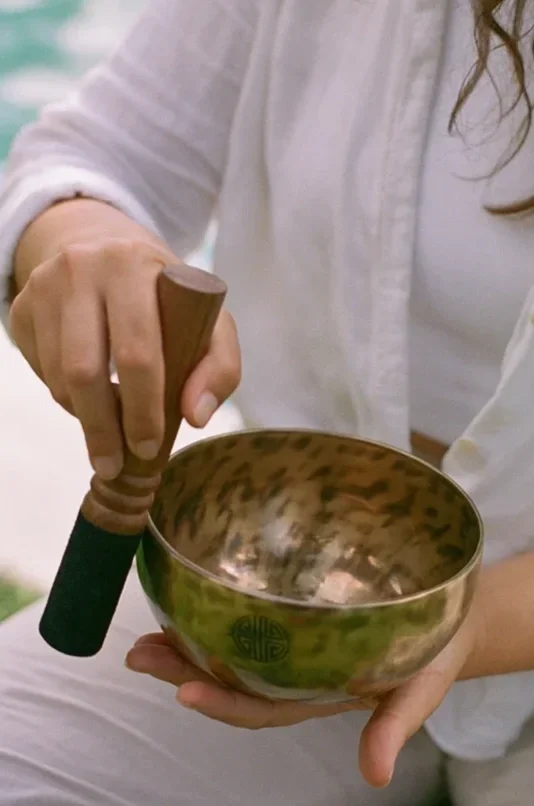 Person holding a singing bowl with a wooden mallet in their right hand, wearing a white shirt for a sound bath in Bali