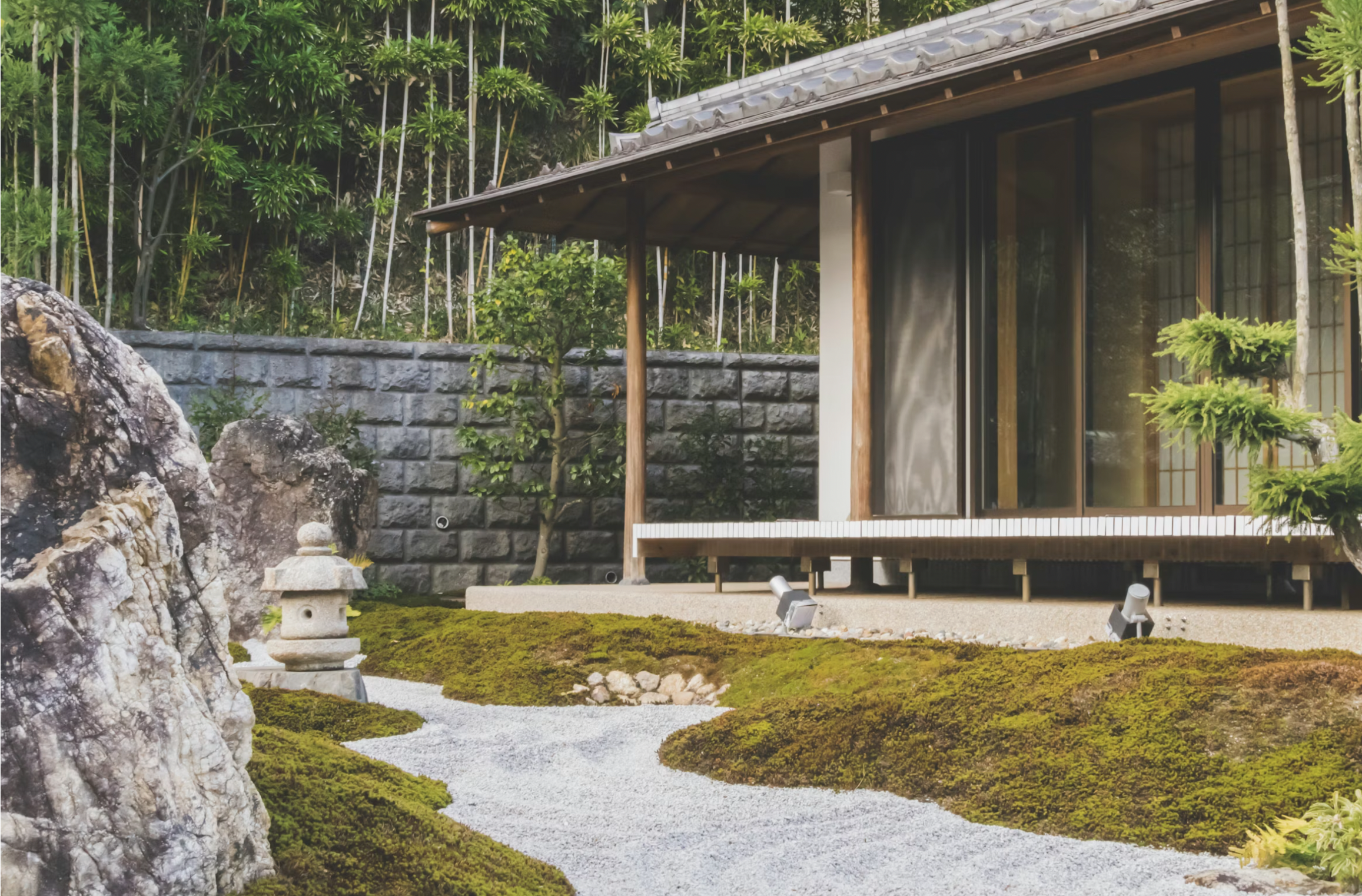 A traditional Japanese Zen garden with a gravel pathway, moss-covered mounds, a stone lantern, and a building with wooden and glass sliding doors, surrounded by lush greenery and a stone wall.