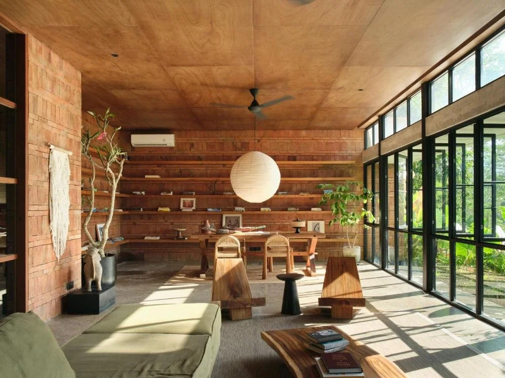 A modern living room with exposed brick walls, a wooden ceiling, and large glass windows. There is a green sofa, a wooden coffee table with books, and a potted plant near the windows. A hanging paper lantern light fixture is in the center, and wooden shelves line the back wall.