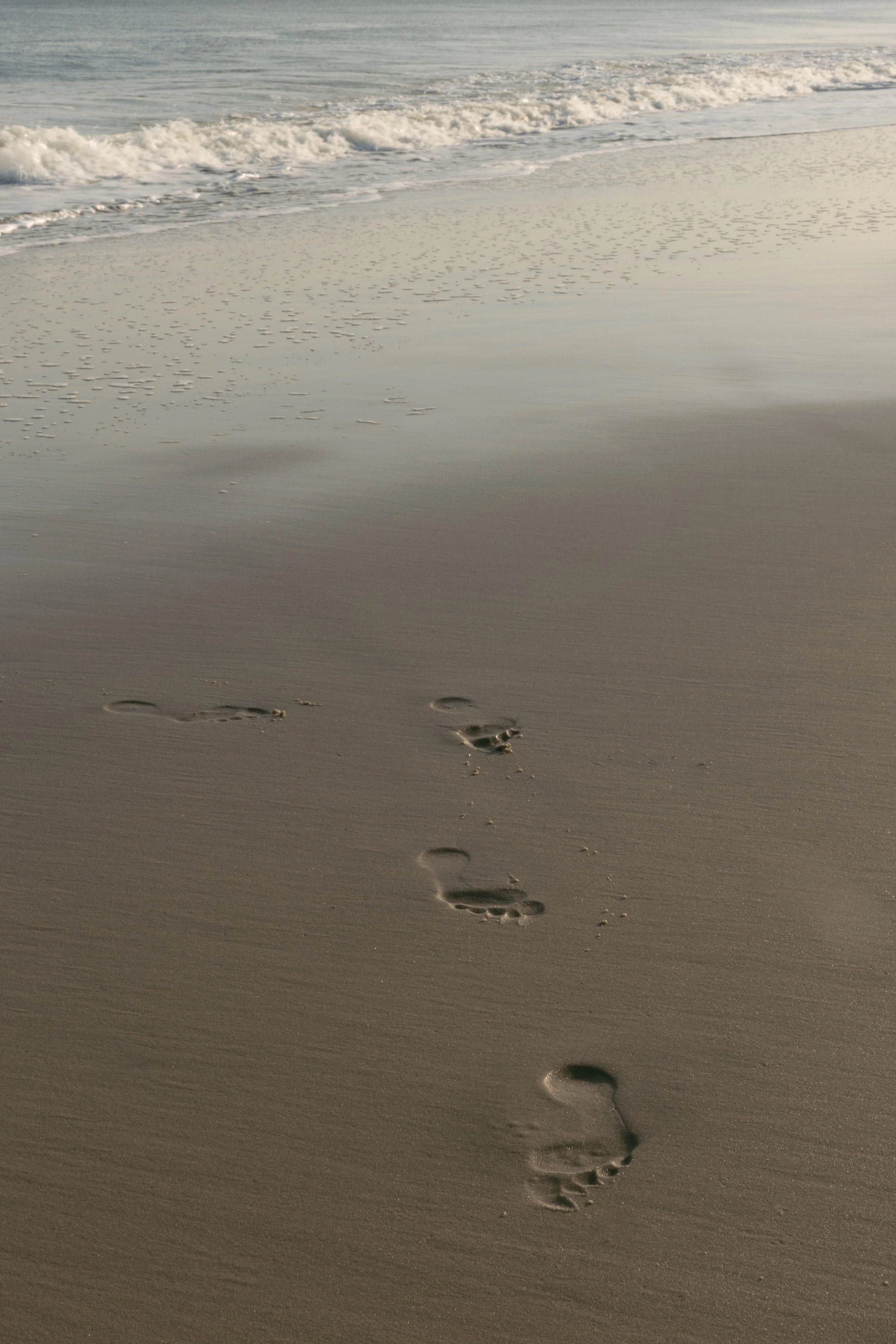 Footprints in the sand leading towards the shoreline of a beach, with gentle waves in the background.