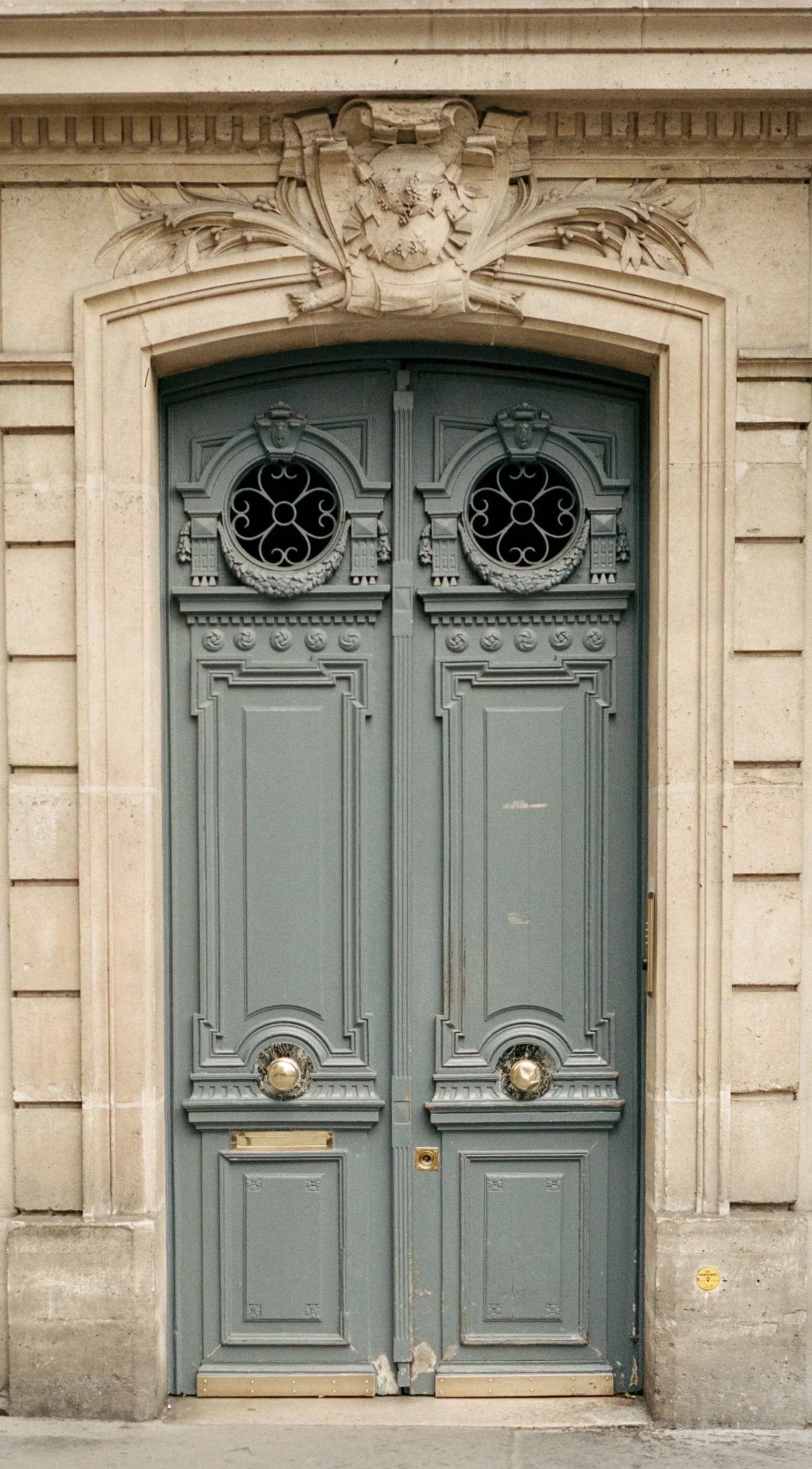 Decorative double door with ornate metalwork, framed by an architectural stone façade with intricate details and a carved emblem above.