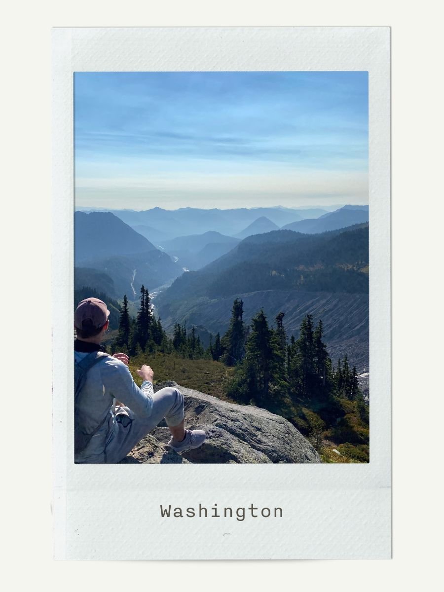 A person sitting on a rock overlooking a mountainous landscape in Washington, with layers of mountains and a cloudy sky in the background.