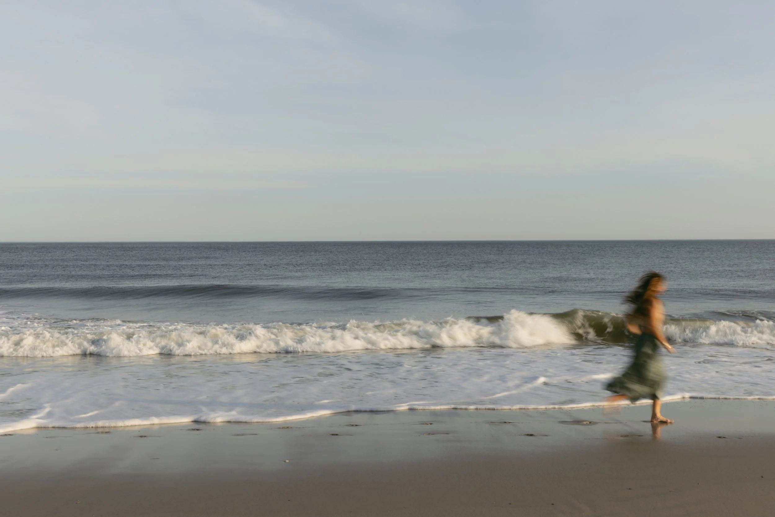 A woman walking along the shoreline at the beach with waves crashing on the sand and a clear sky above.