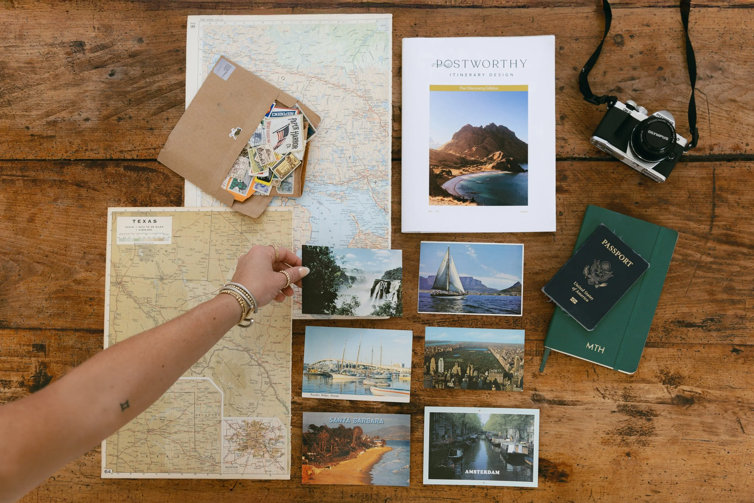 Travel planning setup on wooden table with maps, postcards, a camera, passport, notebooks, and photographs of various scenic locations.