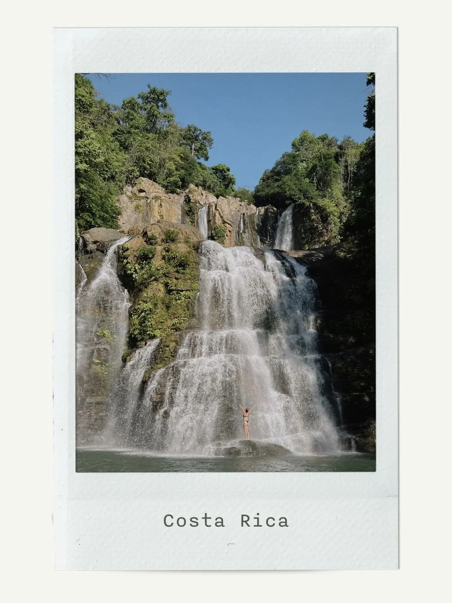 Waterfall in Costa Rica with a person standing at the base, surrounded by lush green trees under a clear blue sky.