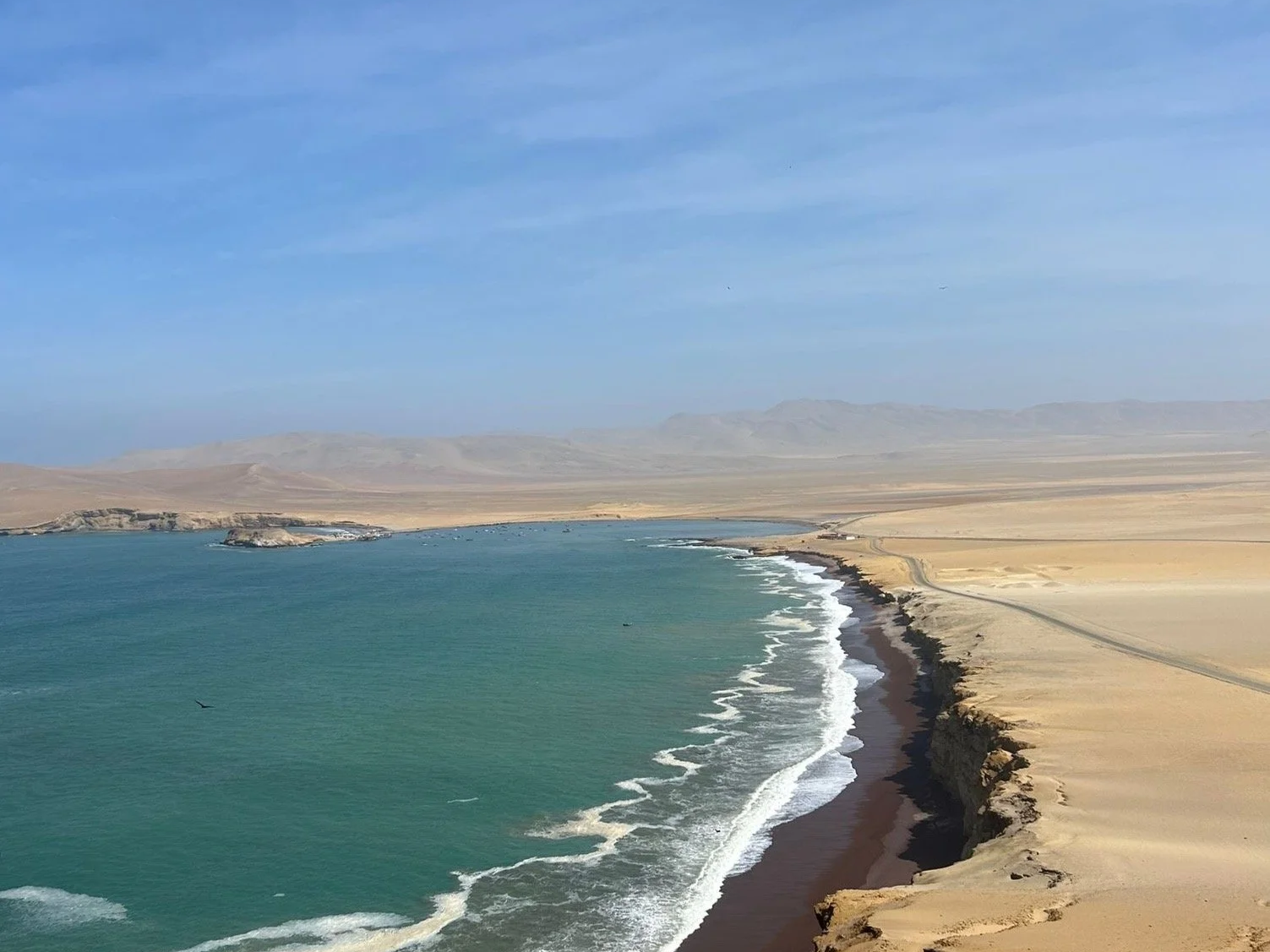 A coastal landscape showing a sandy beach with cliffs, the ocean with waves, and distant mountains under a blue sky with some clouds.