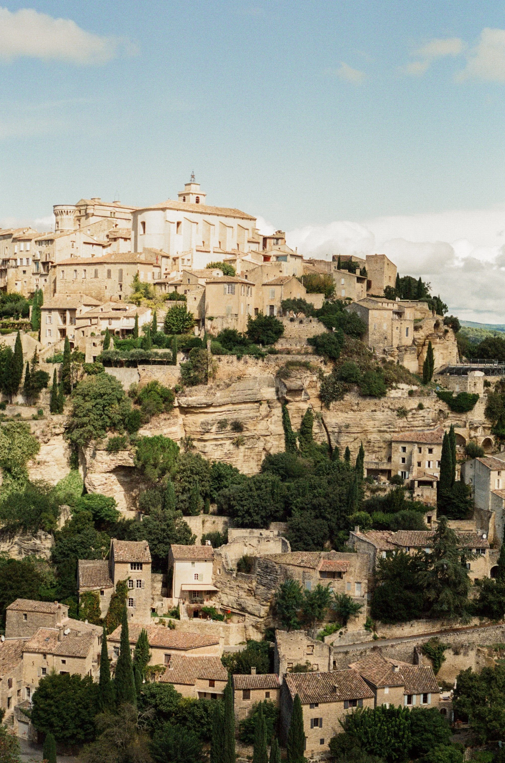 A hillside town with stone buildings in the French countryside, lush greenery, and cypress trees under a partly cloudy sky.