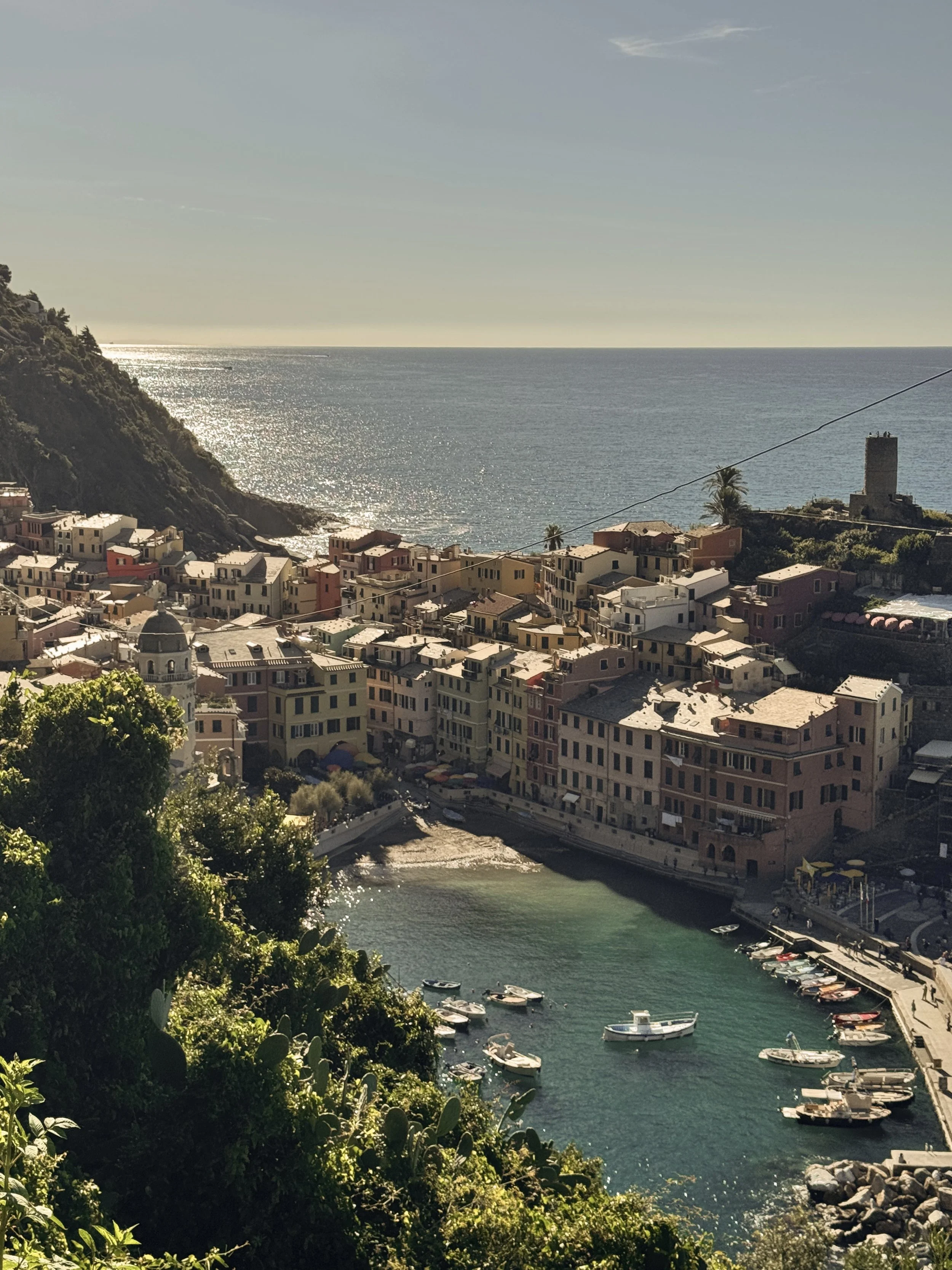 Colorful seaside village with boats in a small harbor, hillside town with ocean view, sunlight reflecting on water, and a distant castle tower.