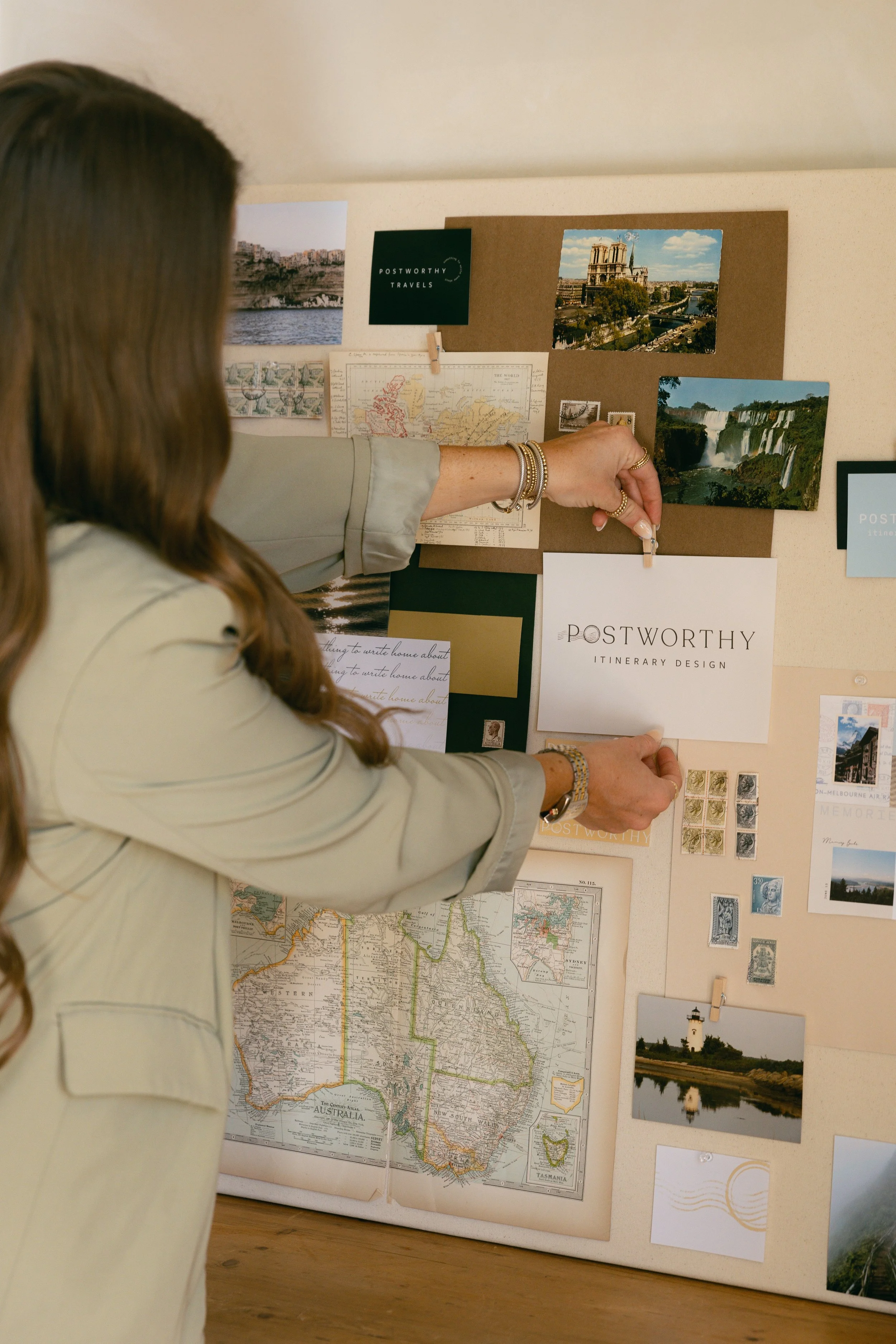 Woman in beige blazer pinning a photo and brochure on a corkboard decorated with travel photos, maps, and stamps.