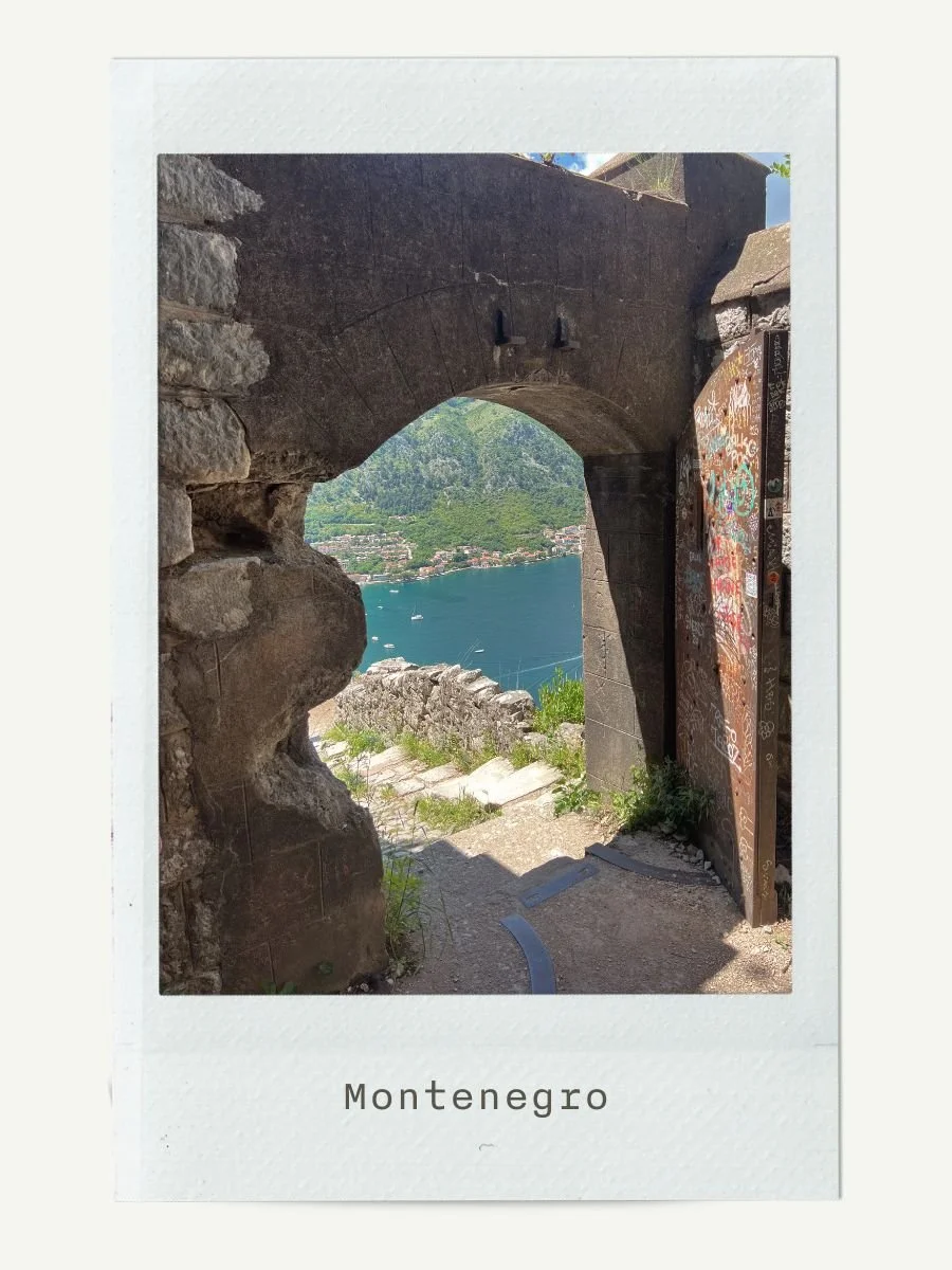 View through a stone archway showing a port with boats on a lake surrounded by green mountains in Montenegro.