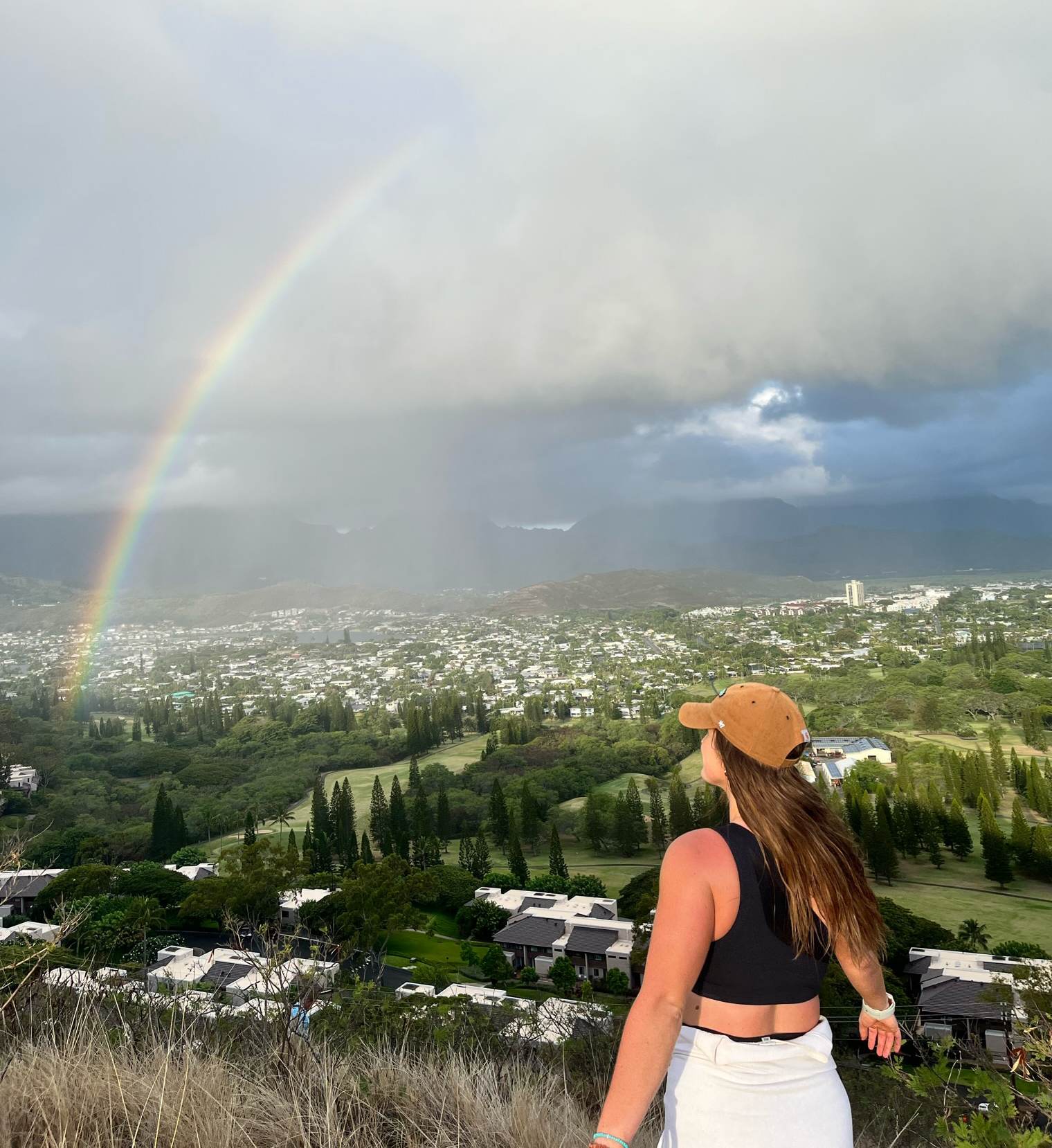 A woman wearing a tan baseball cap, black sleeveless top, and white skirt standing outdoors on a hilltop with a rainbow and a cloudy sky in the background, overlooking a cityscape with greenery.