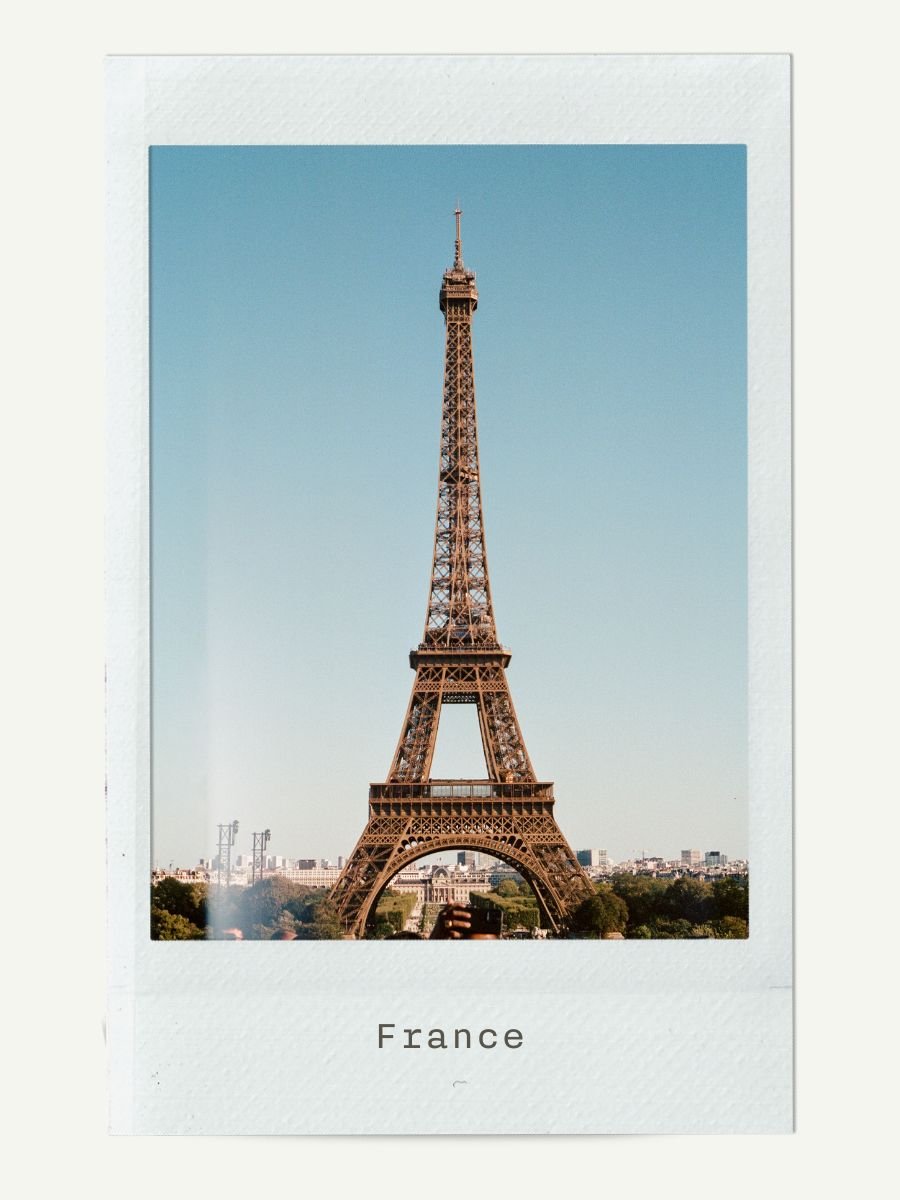 Photograph of the Eiffel Tower in France, with a clear blue sky background and a subtle reflection of a person taking the photo.