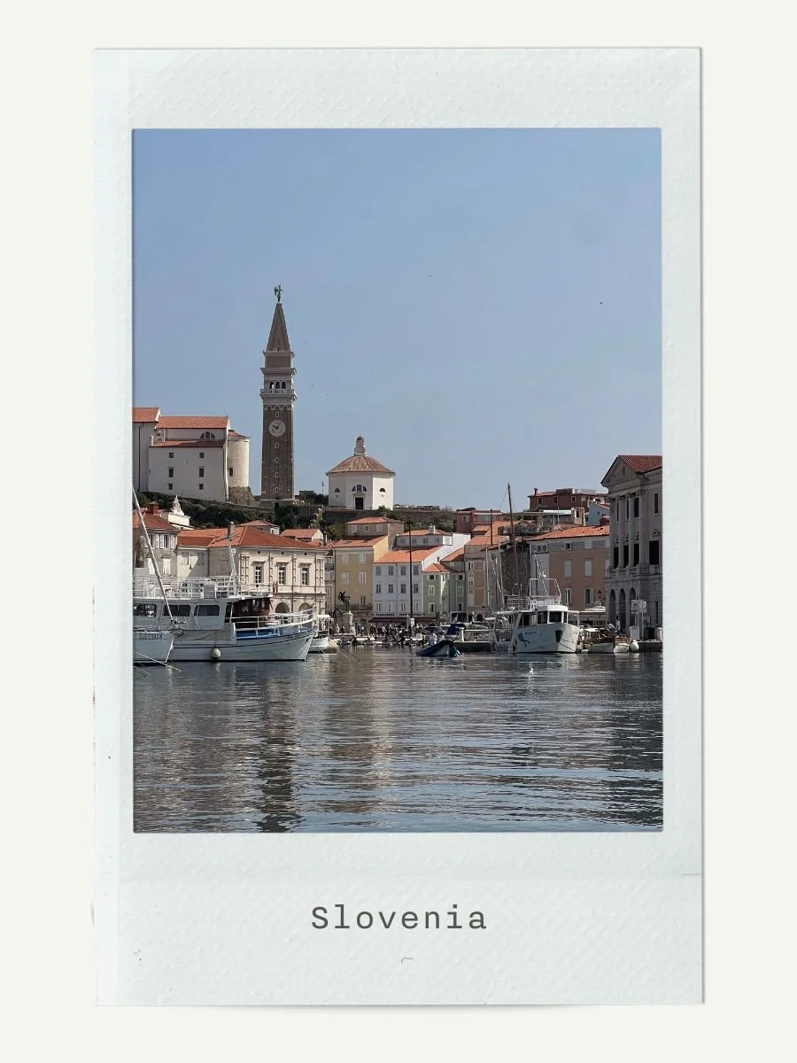 View of a Slovenian harbor with boats anchored in calm water, colorful buildings along the waterfront, and a clock tower on a hill under a clear blue sky.