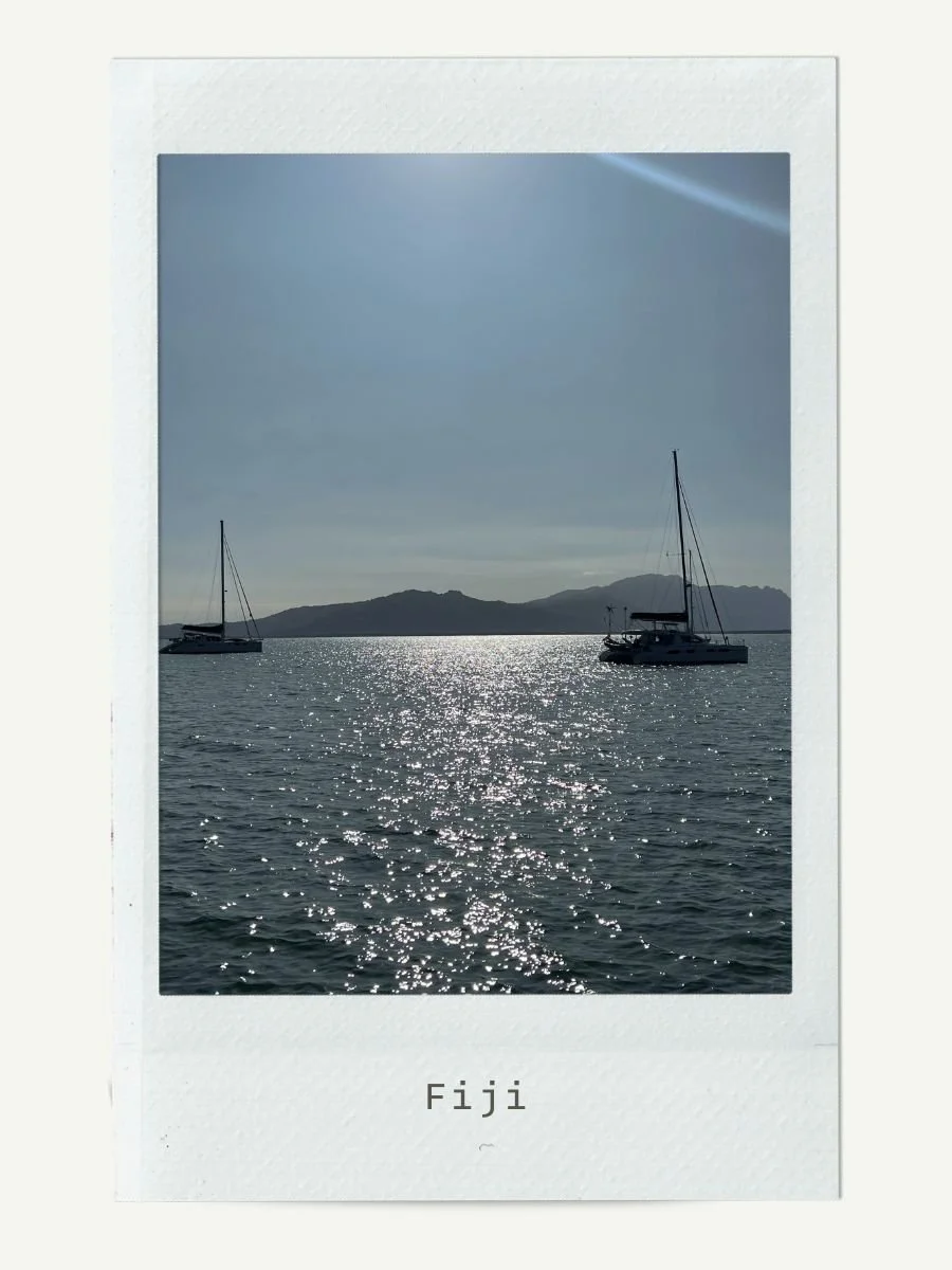 A photo of two sailboats floating on the water near Fiji, with mountains in the background and the sun reflecting on the water.