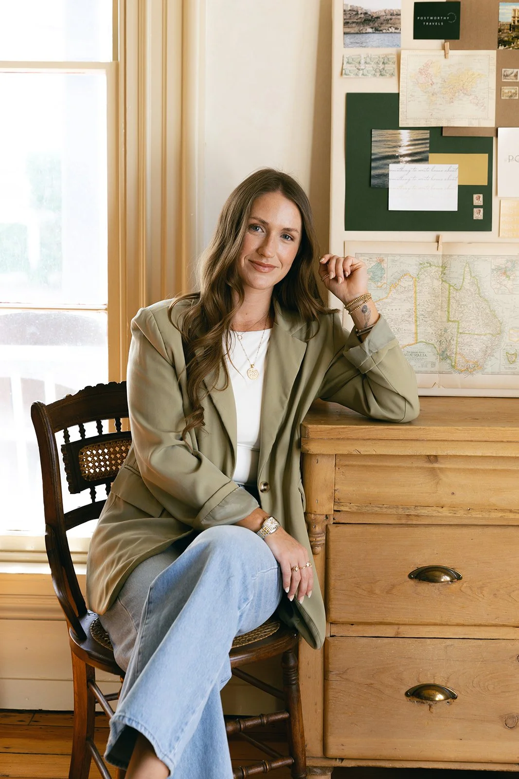 A woman sitting on a wooden chair next to a wooden dresser with maps and postcards on the wall behind her.