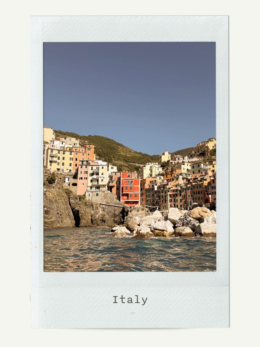 Colorful buildings on a hillside in Italy overlooking the water with rocks in the foreground and a clear blue sky.