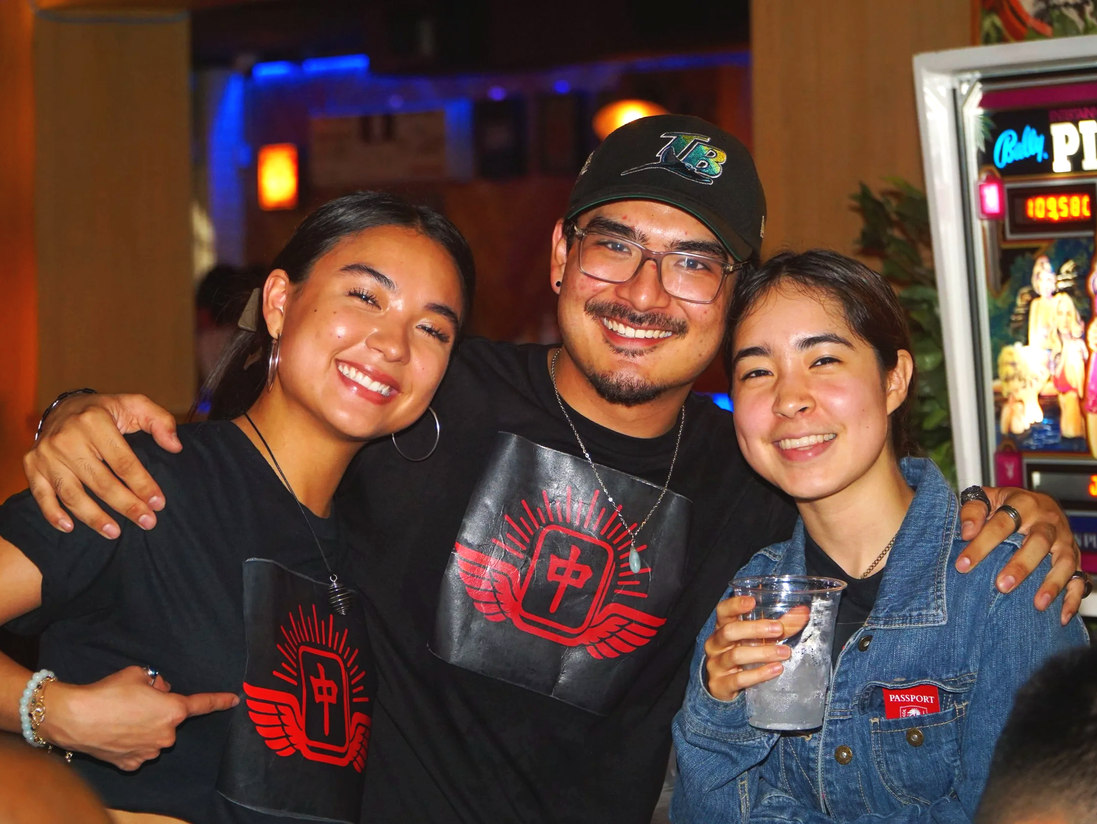 Three young people smiling and hugging in a bar or pub, with a poker machine in the background. They are wearing casual clothes, with two women holding drinks.