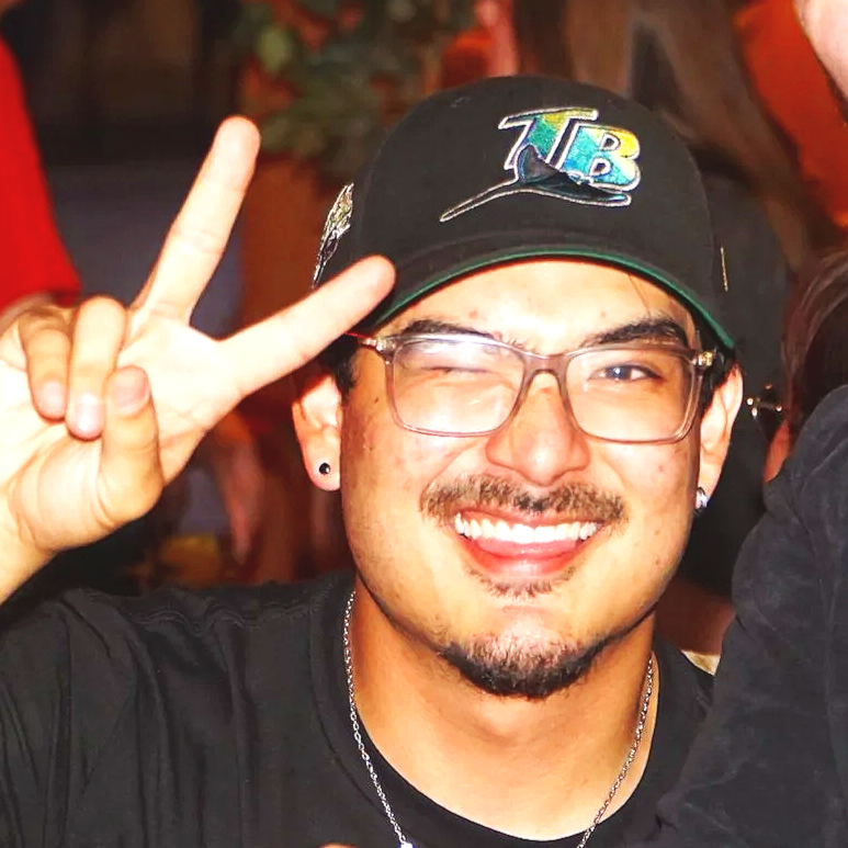 Young man smiling and making a peace sign with his right hand, wearing glasses, a black cap with a colorful logo, and a black shirt in a social setting.