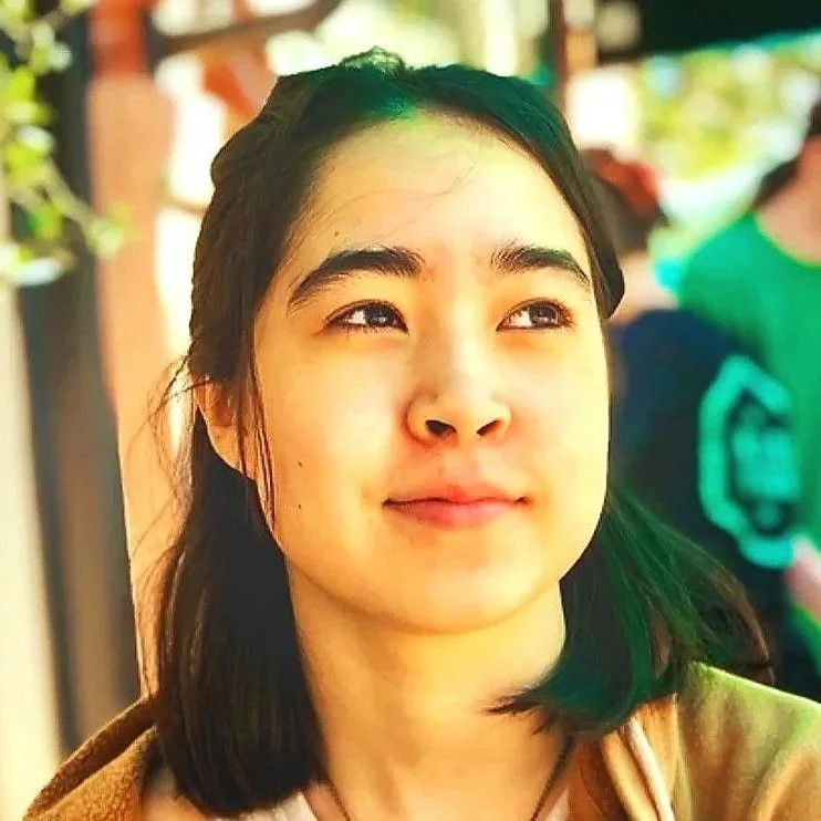 A young woman with dark hair looking thoughtfully to the side, outdoors on a sunny day.