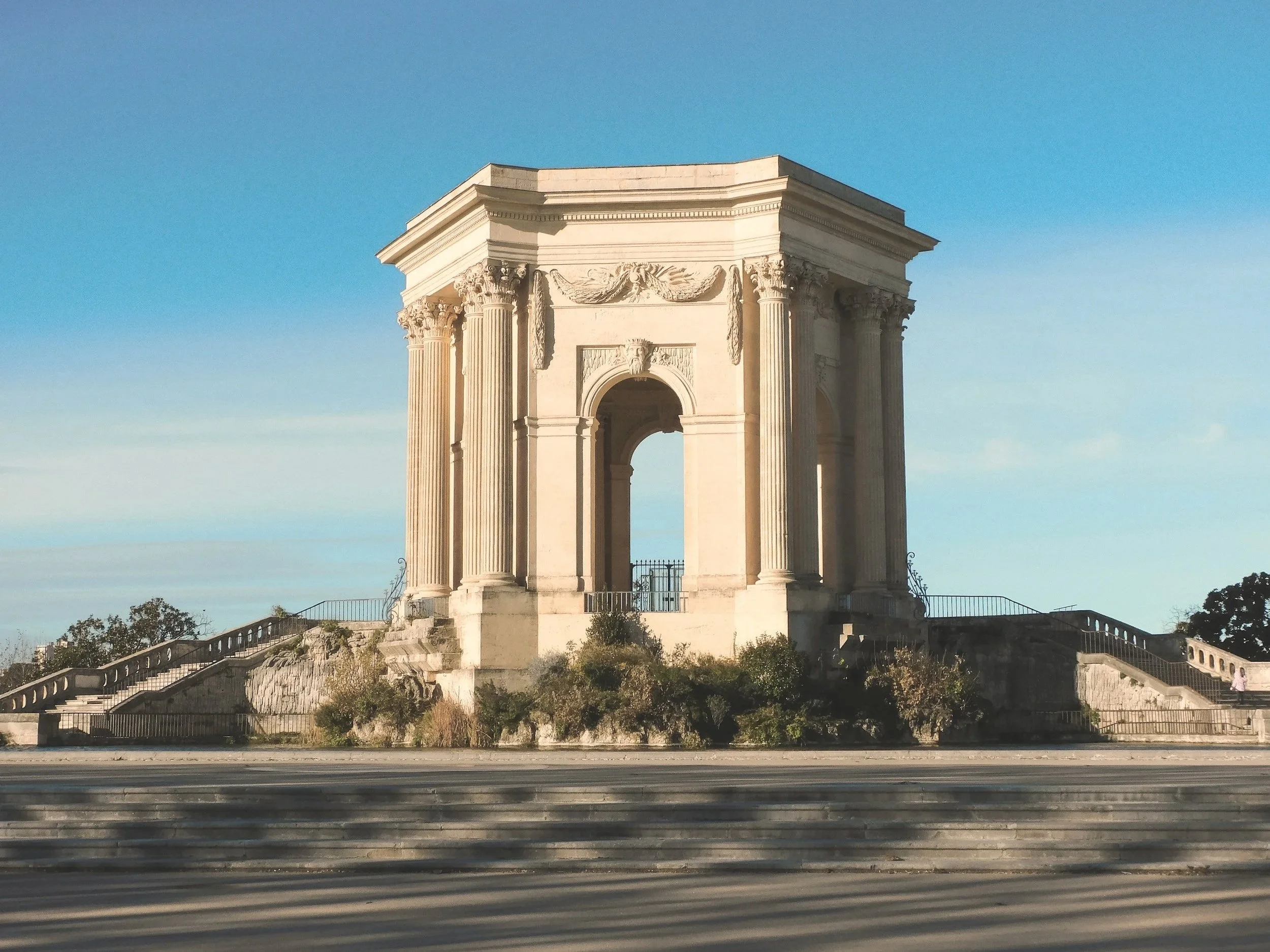 Un monument historique avec des colonnes classiques et une architecture élégante, situé sur une petite colline avec des escaliers menant à l'entrée, sous un ciel bleu clair.