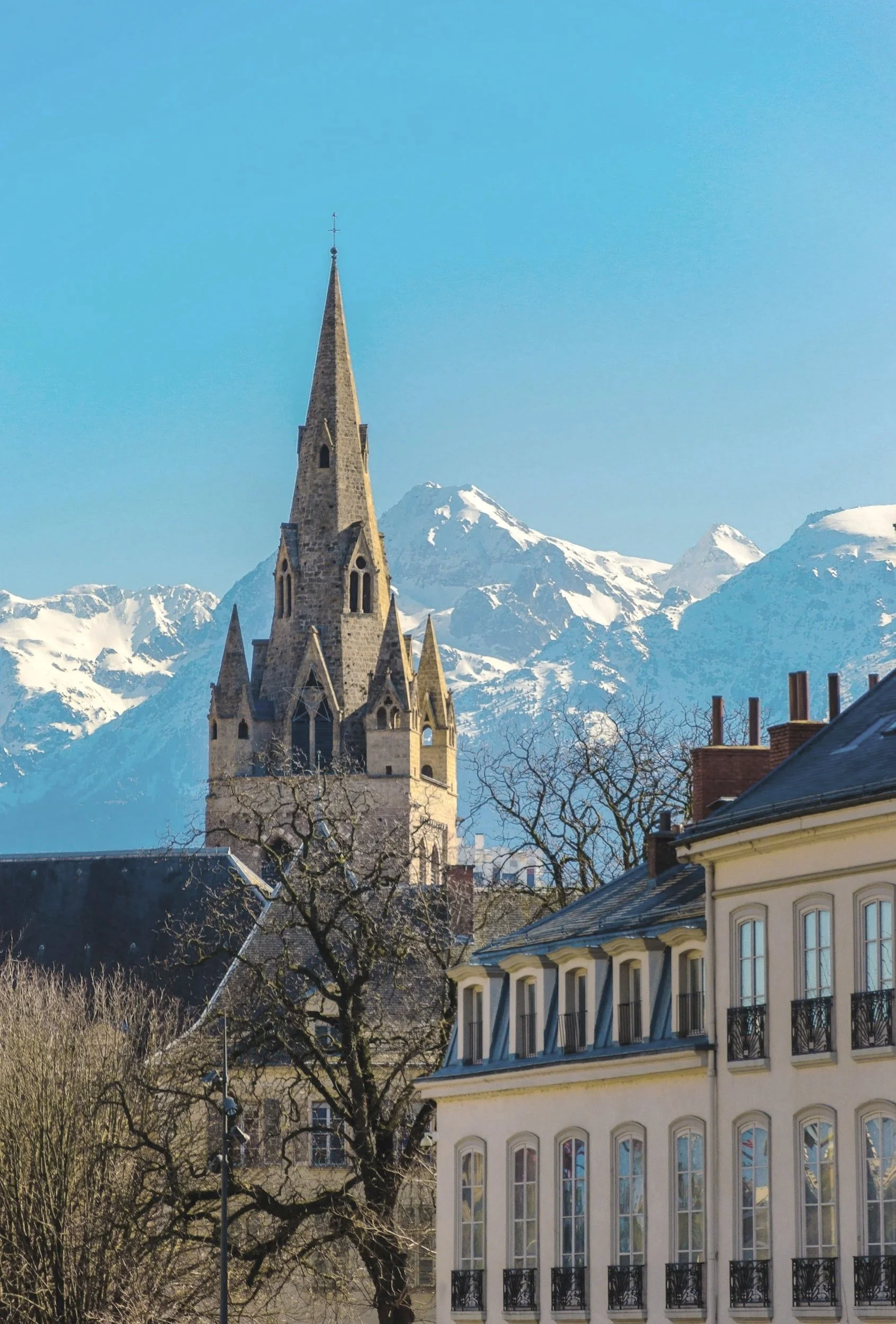 Une église avec une grande tour pointue, des montagnes enneigées en arrière-plan, des bâtiments résidentiels et des arbres sans feuilles.