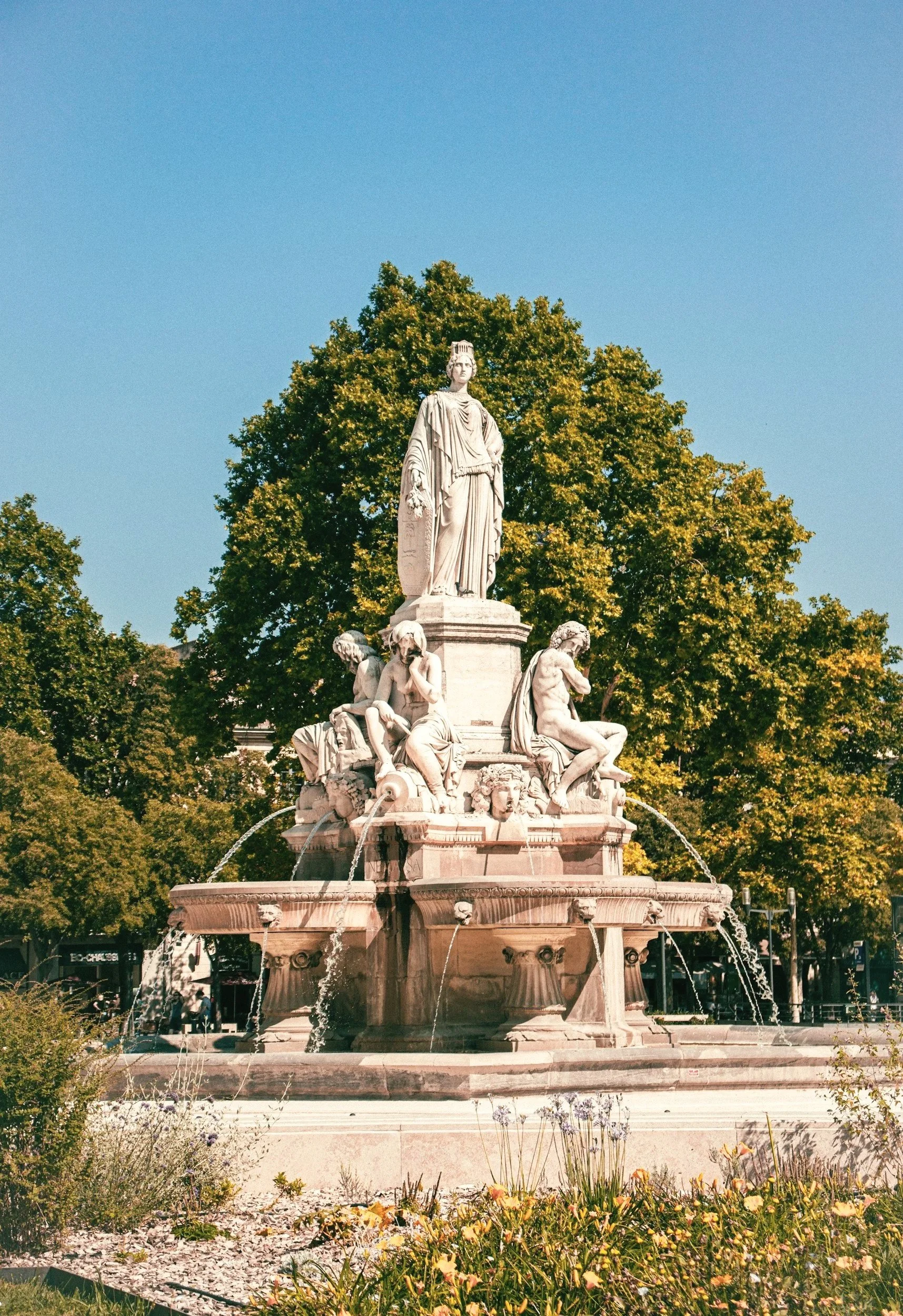Fontaine en pierre avec plusieurs sculptures, située dans un parc avec des arbres verts en arrière-plan, sous un ciel bleu clair.