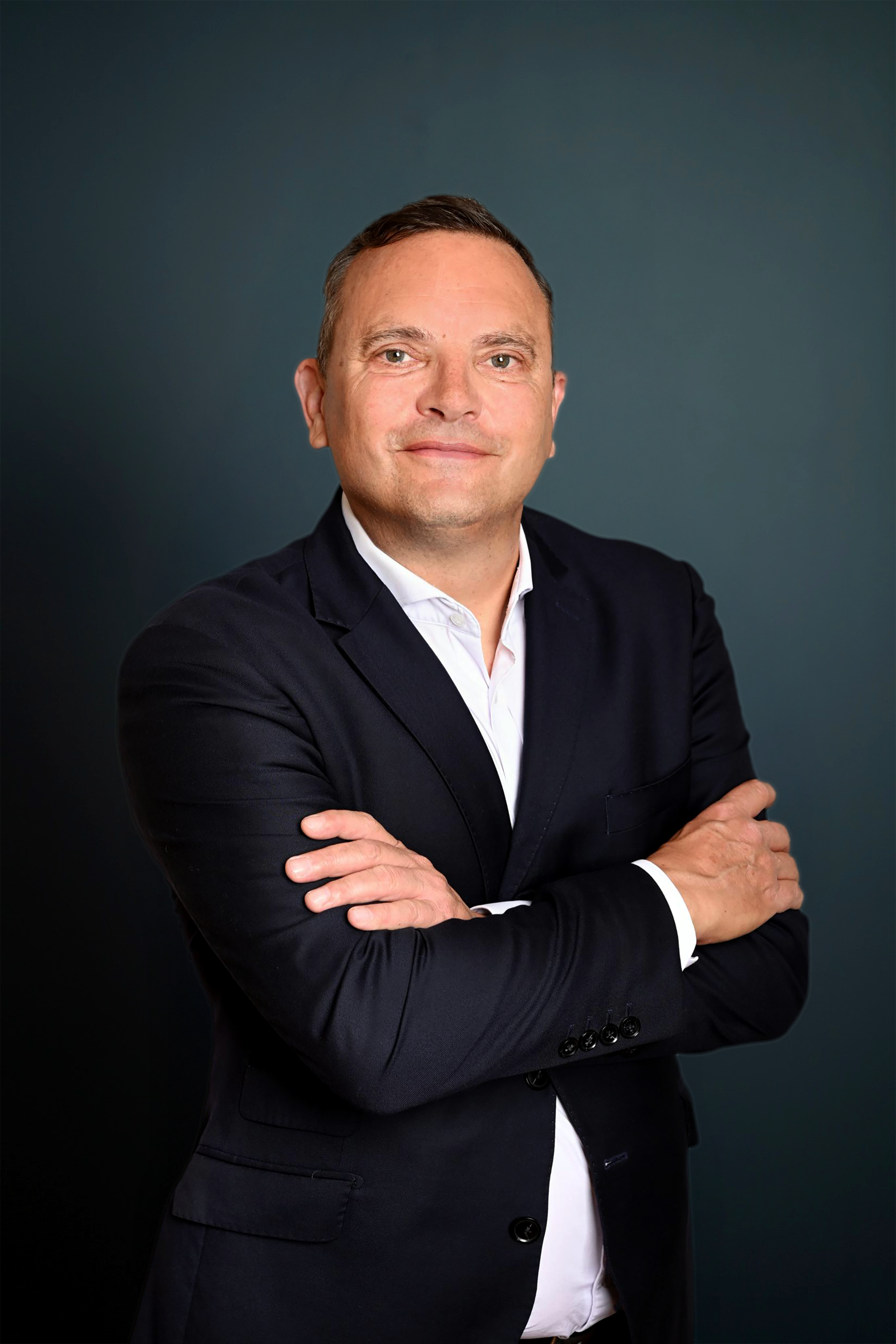 Homme d'affaires portant un costume noir avec une chemise blanche, croisant les bras, souriant devant un fond sombre.