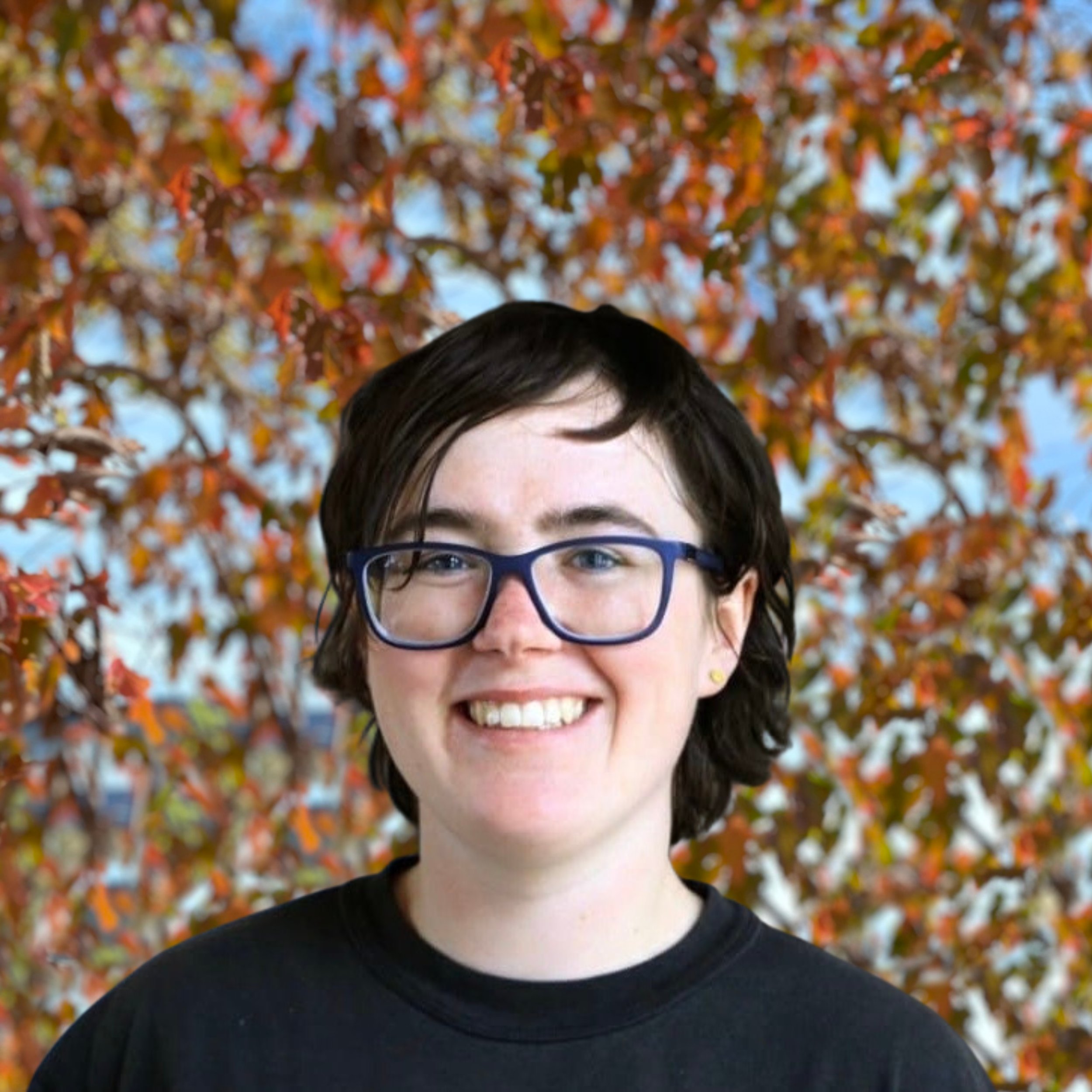 Portrait einer lächelnden Person mit kurzen dunklen Haaren, blaue Brille, schwarze T-Shirt, vor einem Baum mit orange-blättrigem Herbstlaub.