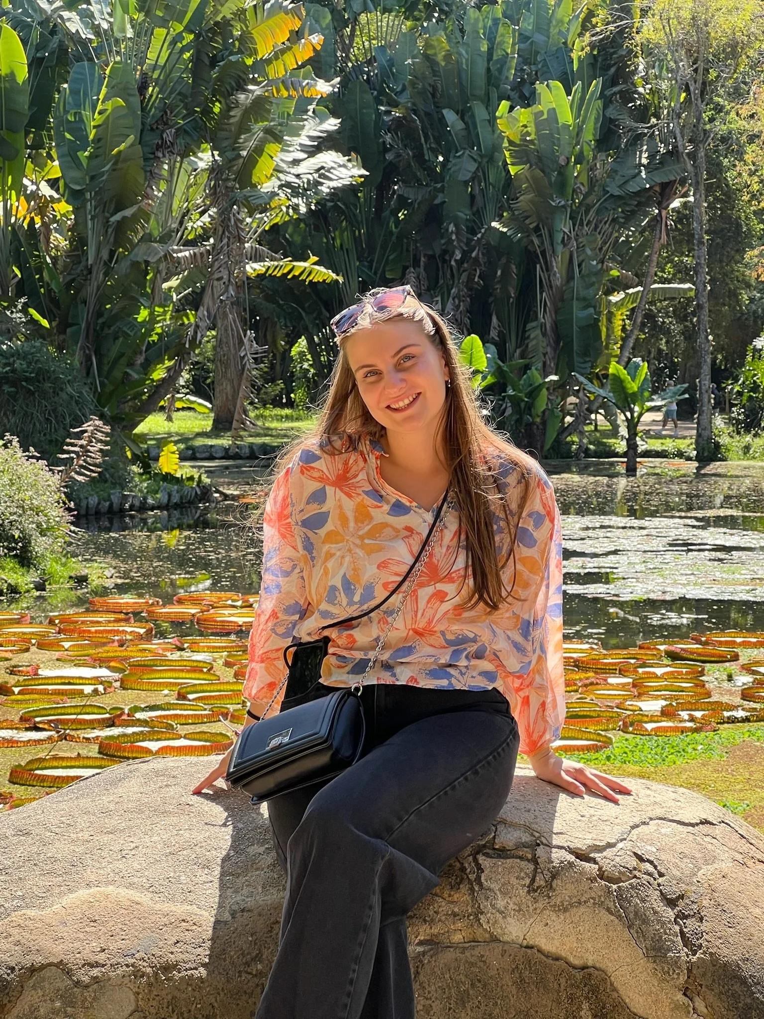 Lola Graff souriante assise sur une roche devant un étang avec des plantes de lotus, entourée de végétation tropicale dense.