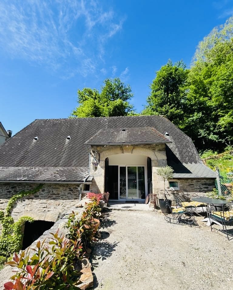 Quaint stone cottage with a steep roof, surrounded by greenery, under a clear blue sky. Outdoor seating area with plants along a gravel path.