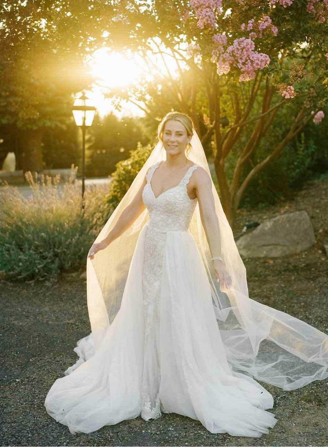 Bride in wedding dress with veil outdoors at sunset, surrounded by trees and flowers.