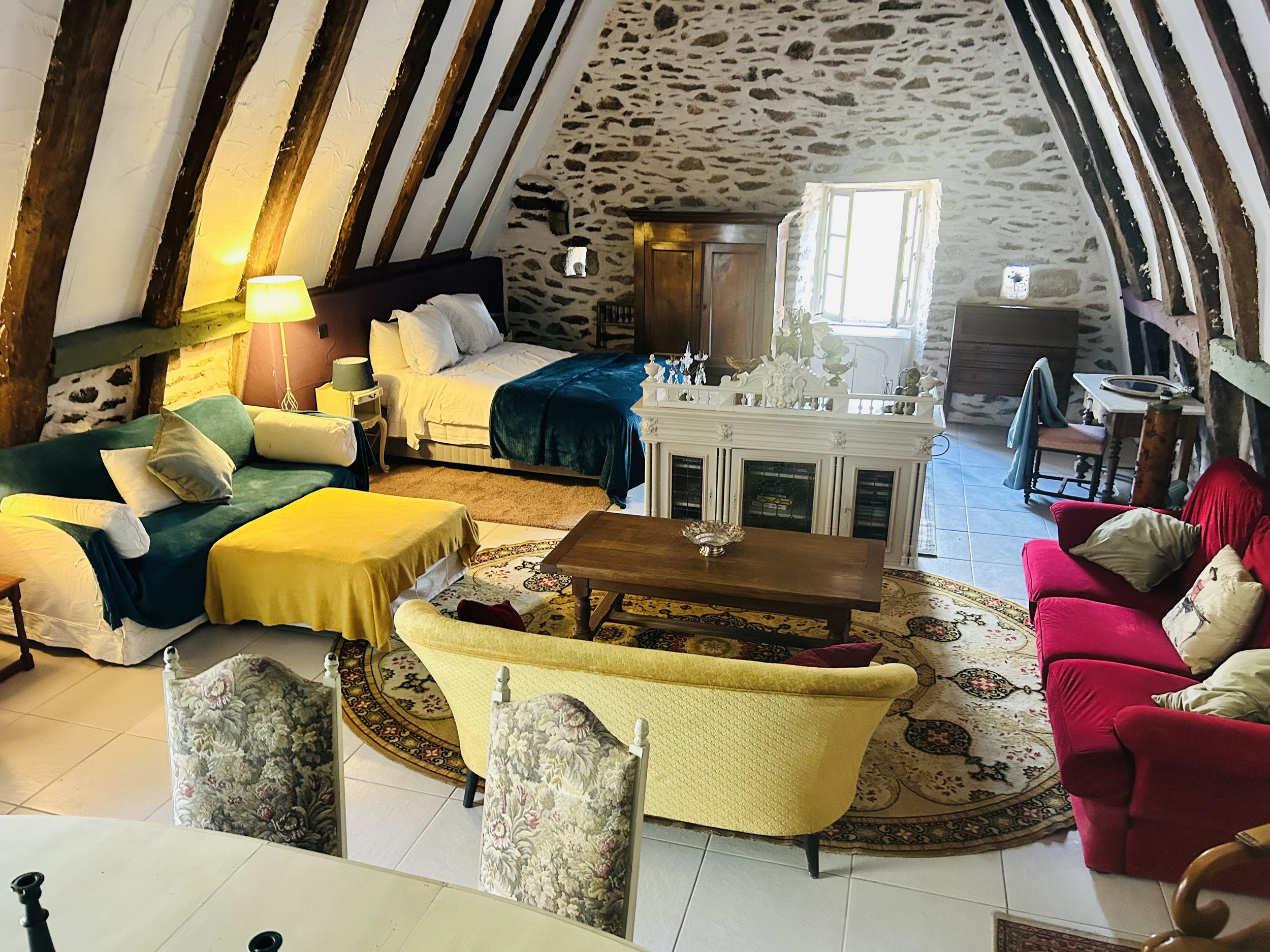 Cozy attic room with exposed wooden beams, stone walls, a double bed, colorful couches, a round rug, and various furniture pieces, including a coffee table and chairs.