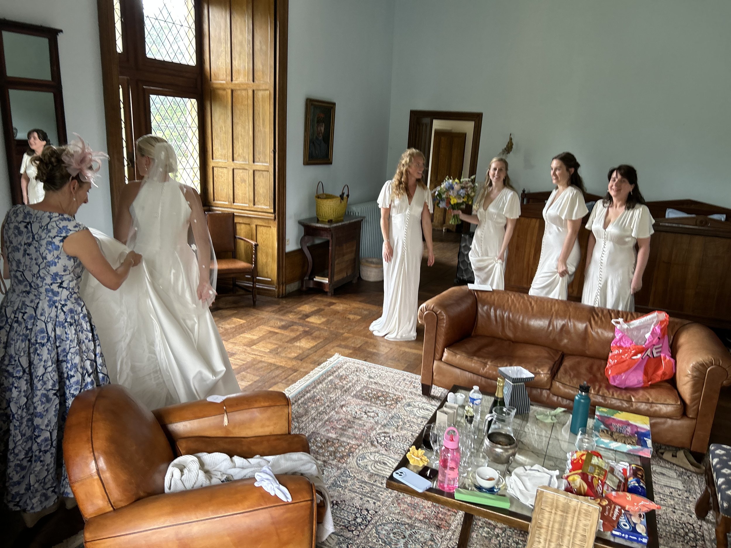 Bridal party in a room preparing for a wedding; a bride is in a white dress and veil being assisted by a woman in a floral dress. Bridesmaids in white dresses are standing nearby, and a living area with a leather sofa and scattered items on a coffee