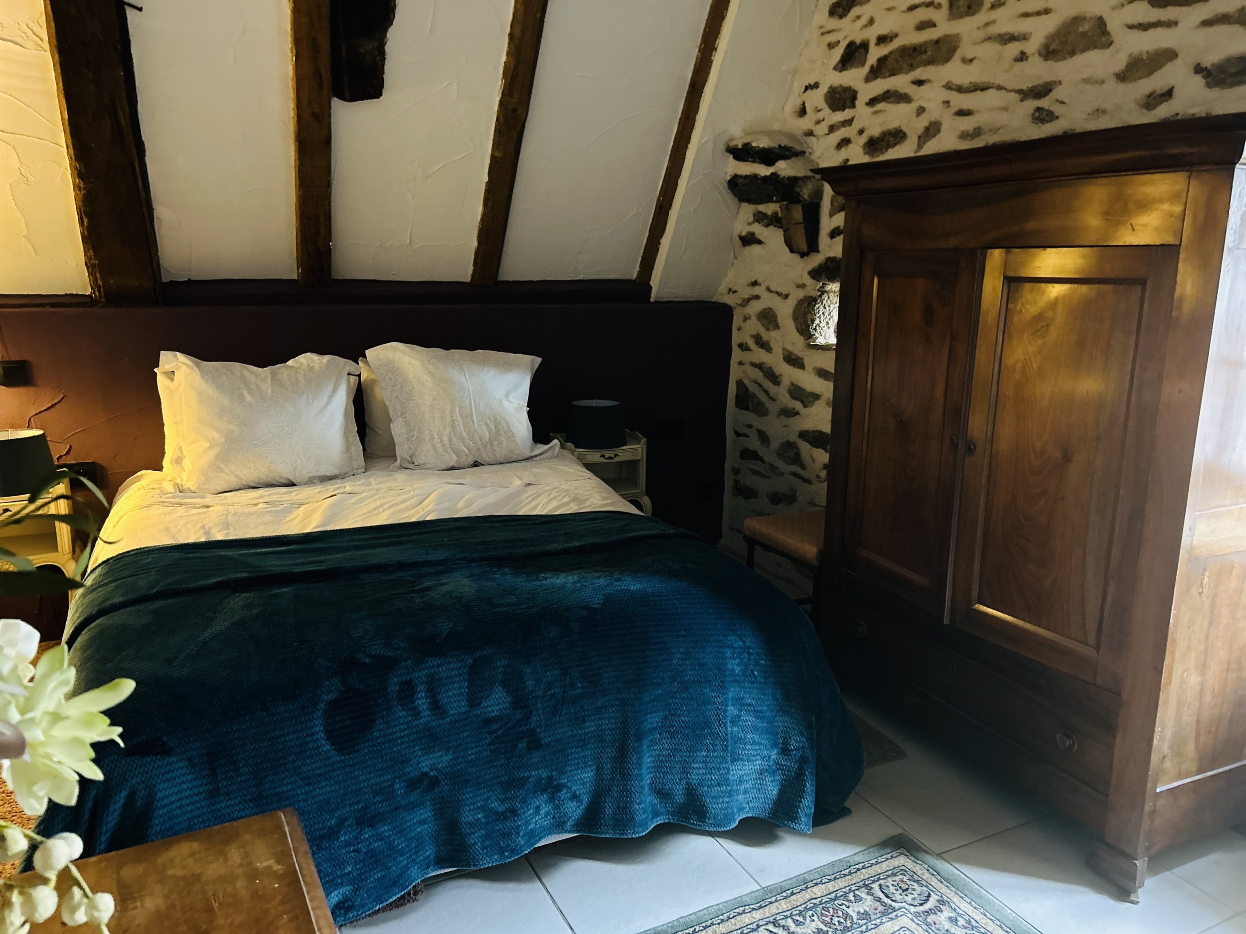 Cozy bedroom with rustic elements, featuring a double bed with white pillows and a dark blue blanket, wooden wardrobe, stone wall, and beams on the ceiling.