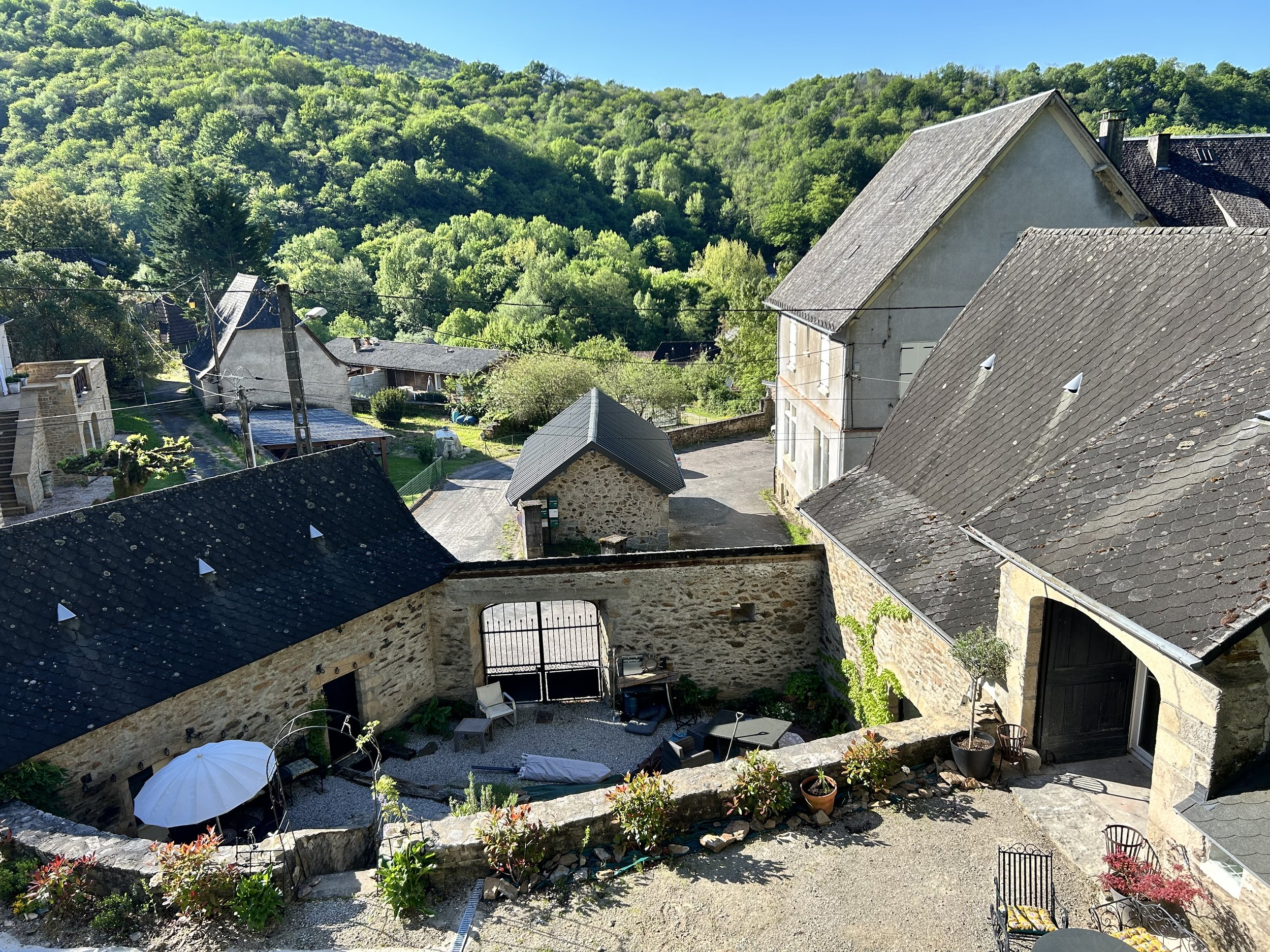 View of a rustic courtyard in a French village with stone buildings, slate roofs, and outdoor seating. Surrounded by lush greenery and rolling hills under a clear blue sky.