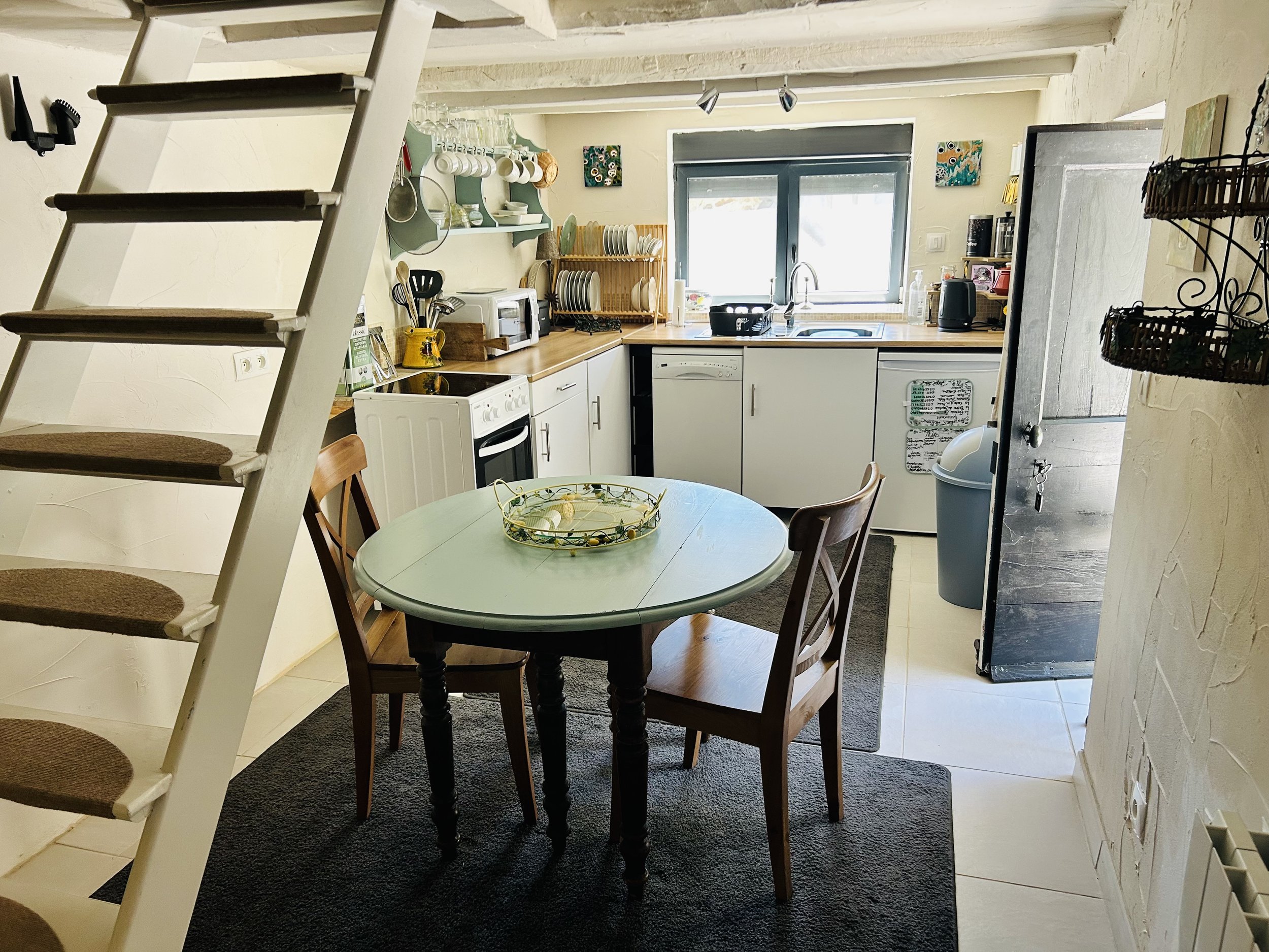 Cozy kitchen interior with a white staircase, round dining table, wooden chairs, and modern appliances. Shelves hold dishes and cups. A door opens to the outside.