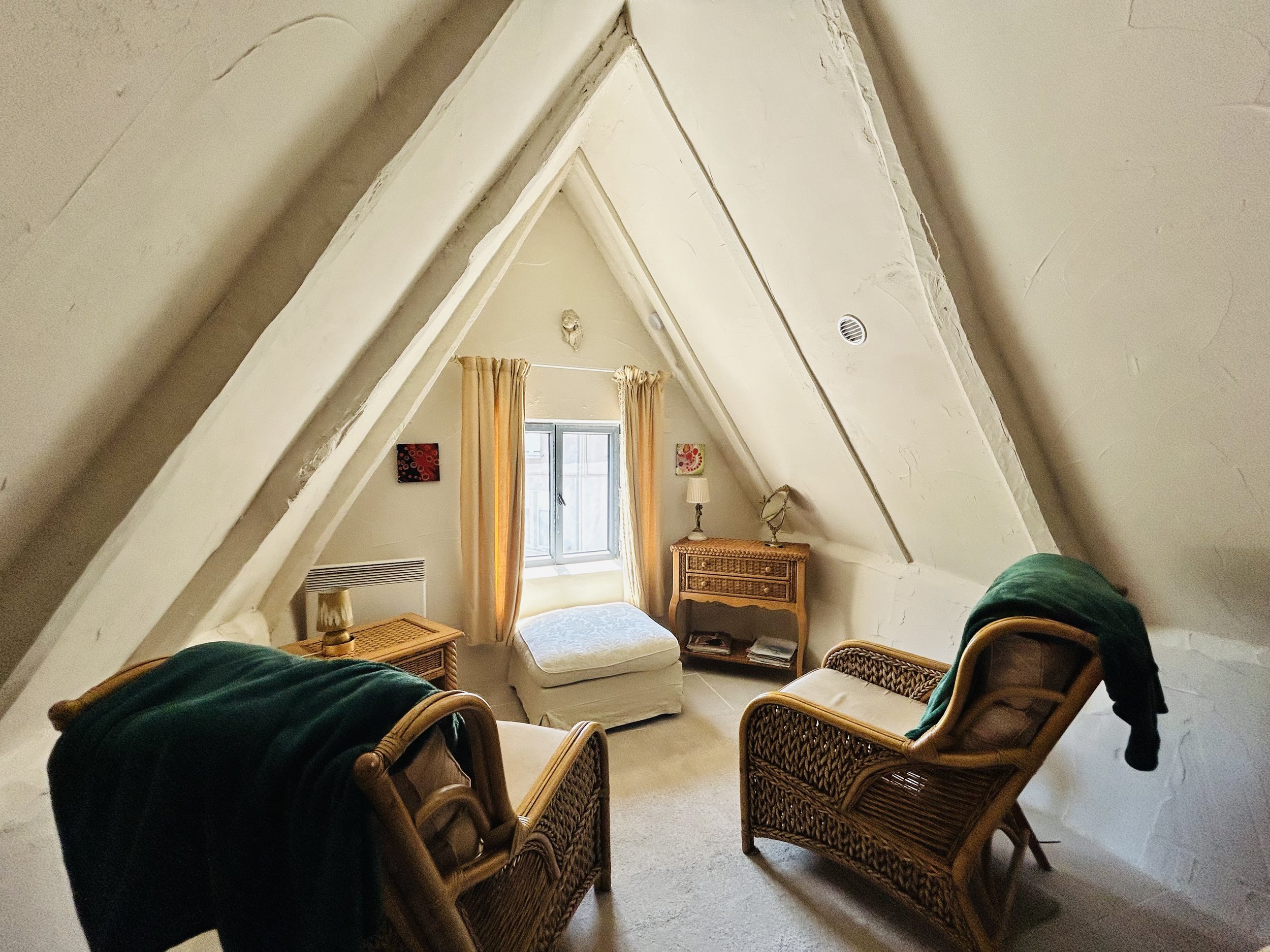 Cozy attic room with slanted ceiling, wicker chairs, green blanket, window with curtains, ottoman, and wicker side tables.