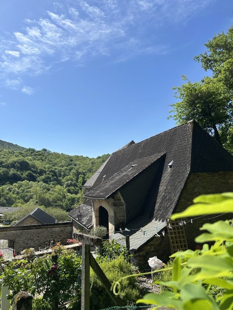 Stone building with a steep roof surrounded by lush greenery and hills under a clear blue sky.