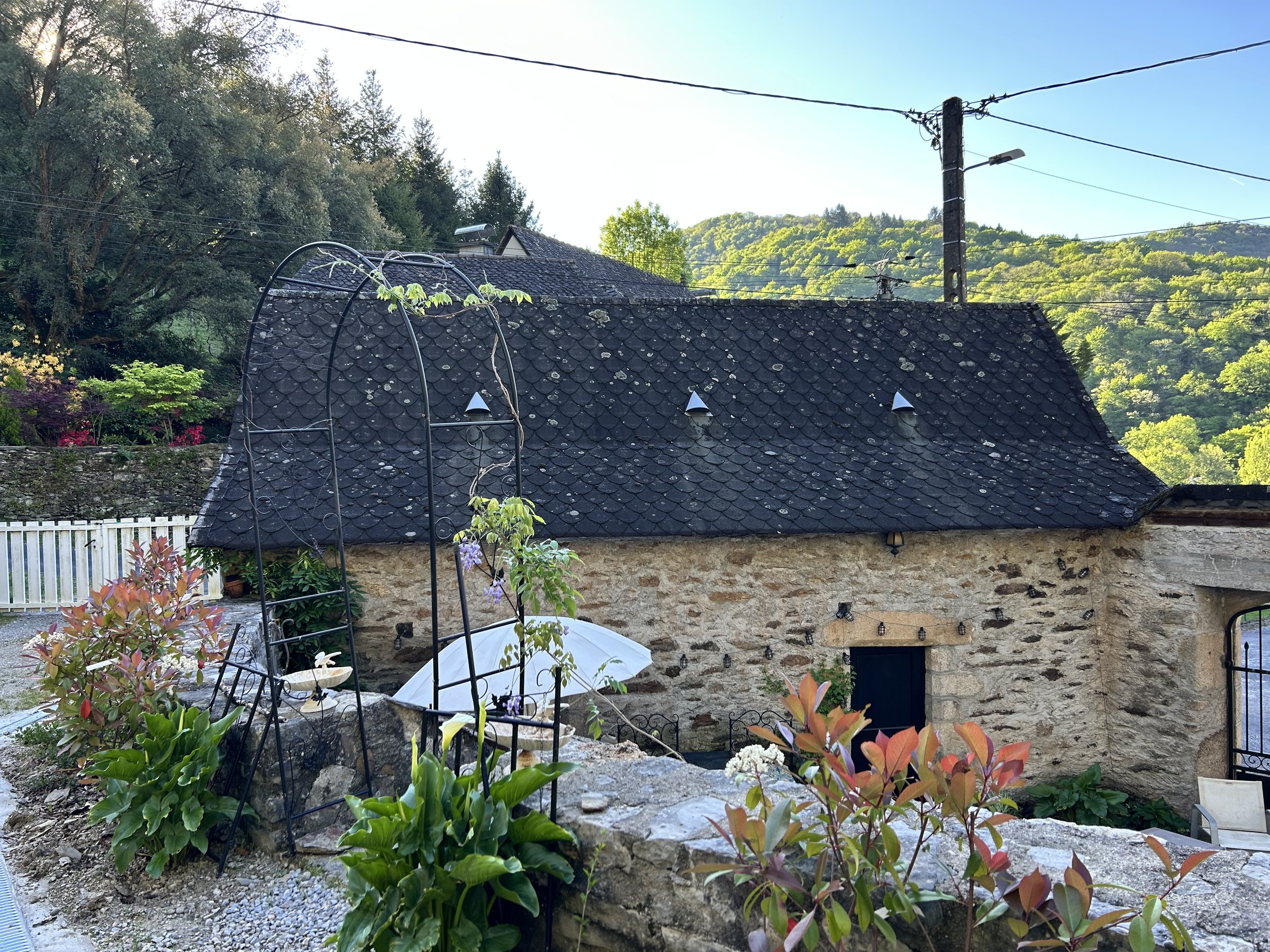 Stone cottage with a dark shingle roof, surrounded by lush greenery and trees, featuring a wrought iron arch and plants in the foreground.