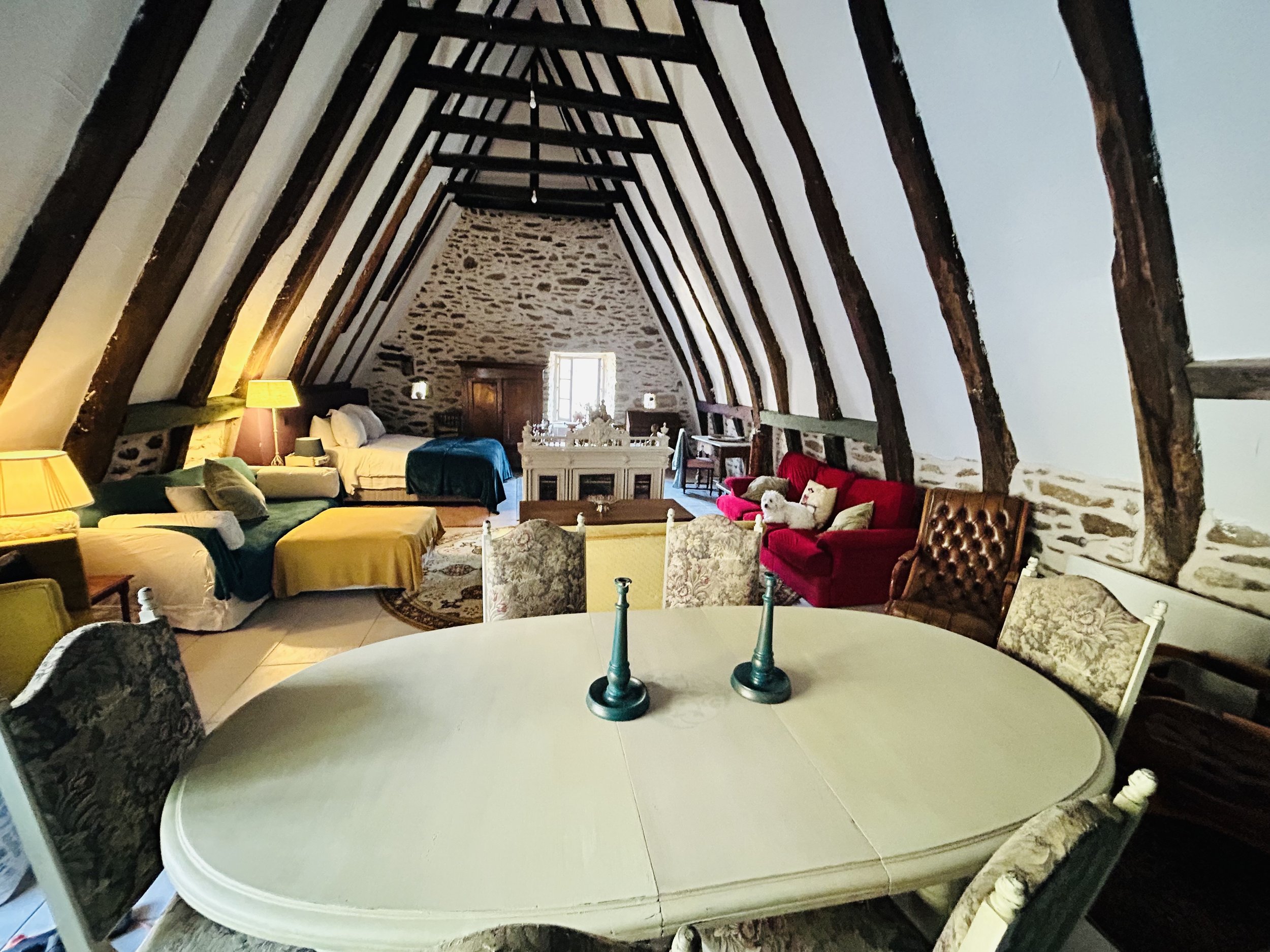 Cozy rustic attic room with exposed wooden beams, dining table, red sofa, armchair, beds, and soft lighting.