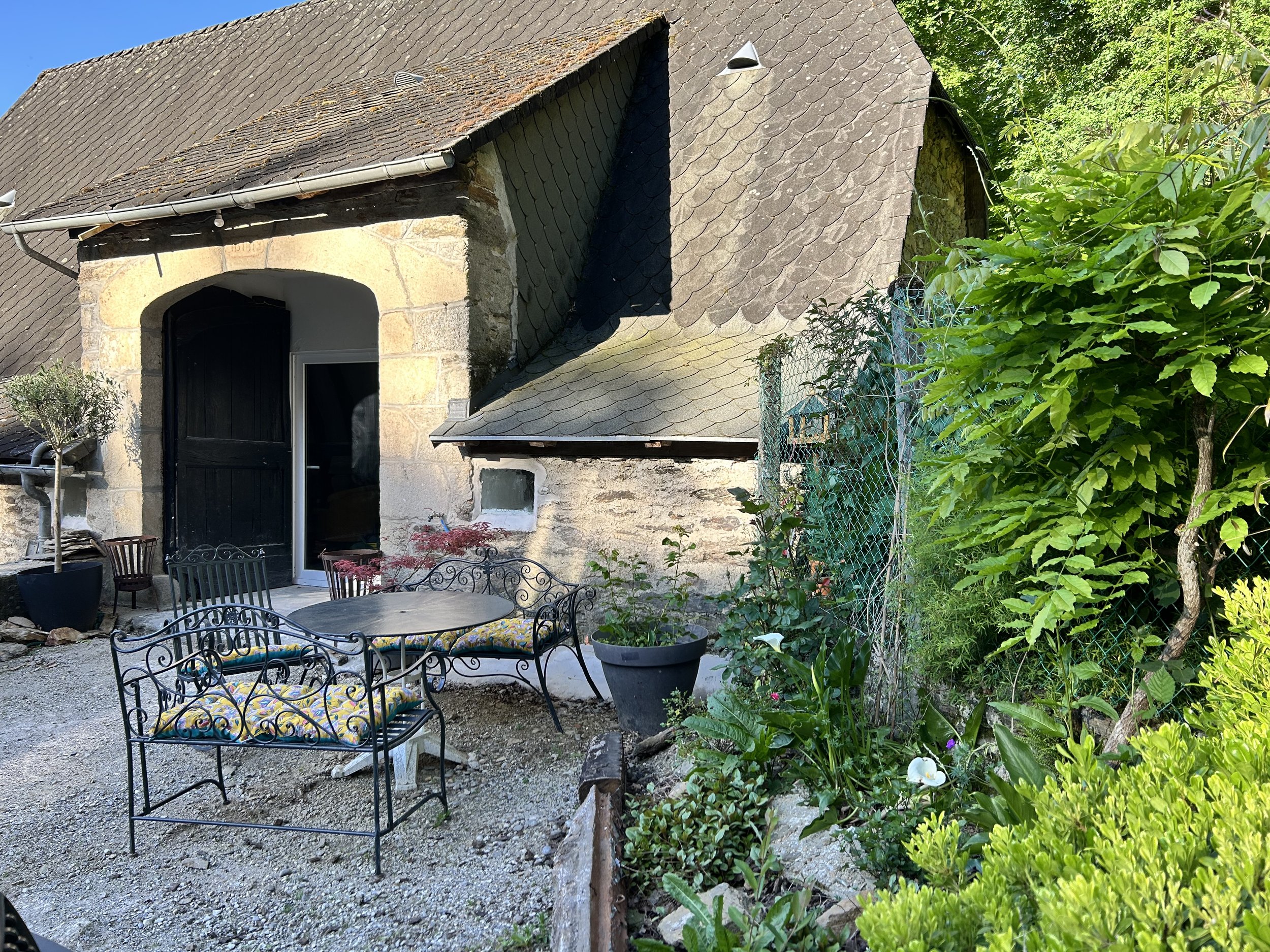Stone cottage with a sloped tile roof, surrounded by greenery. Outside, there's wrought iron patio furniture with cushions and potted plants on a gravel area.