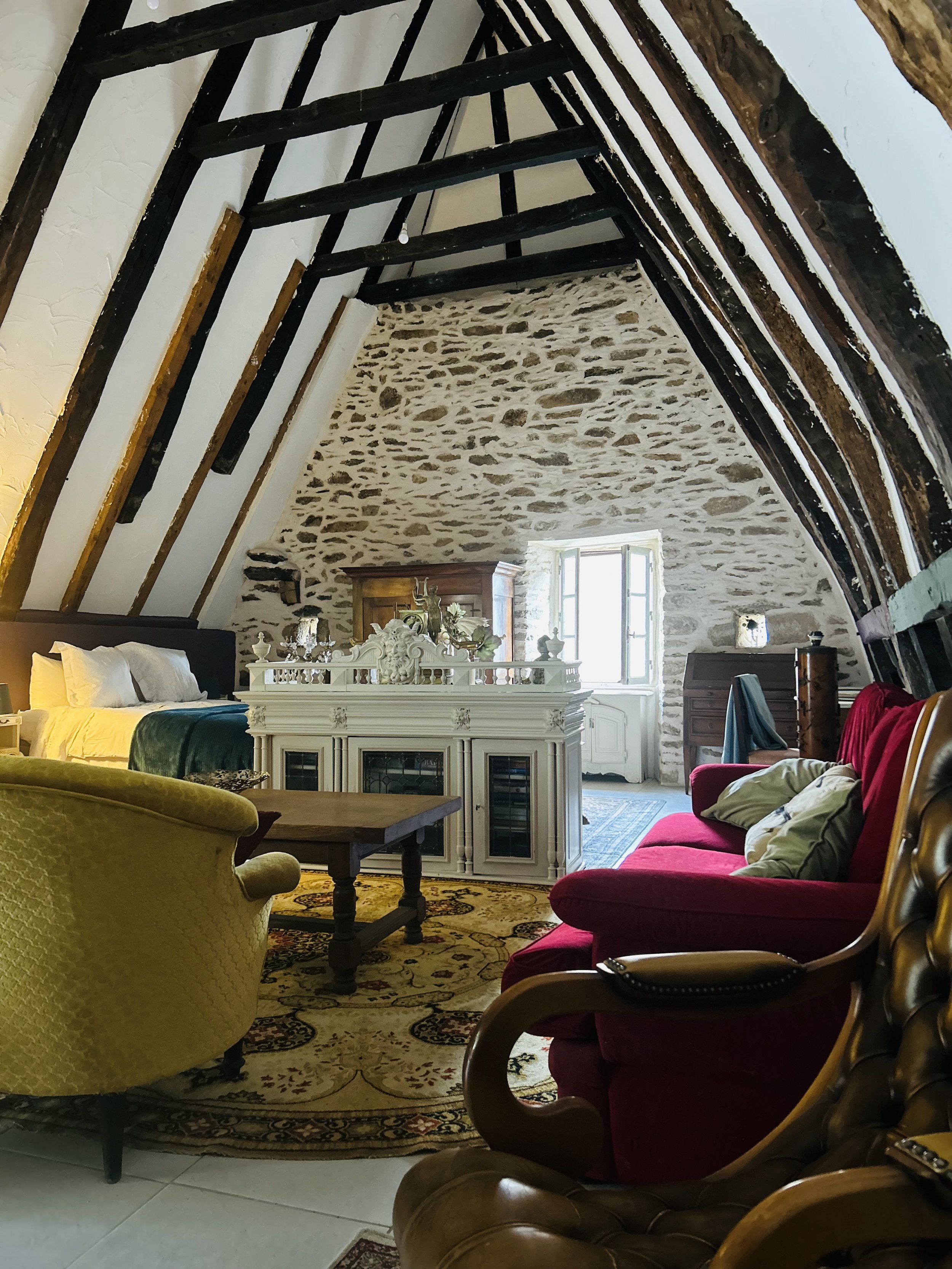 Interior of rustic attic bedroom with exposed beams, stone walls, and vintage furniture including a red sofa, yellow armchair, and wooden chairs. A bed with white linens is visible, and a decorative fireplace with ornaments stands near a small window