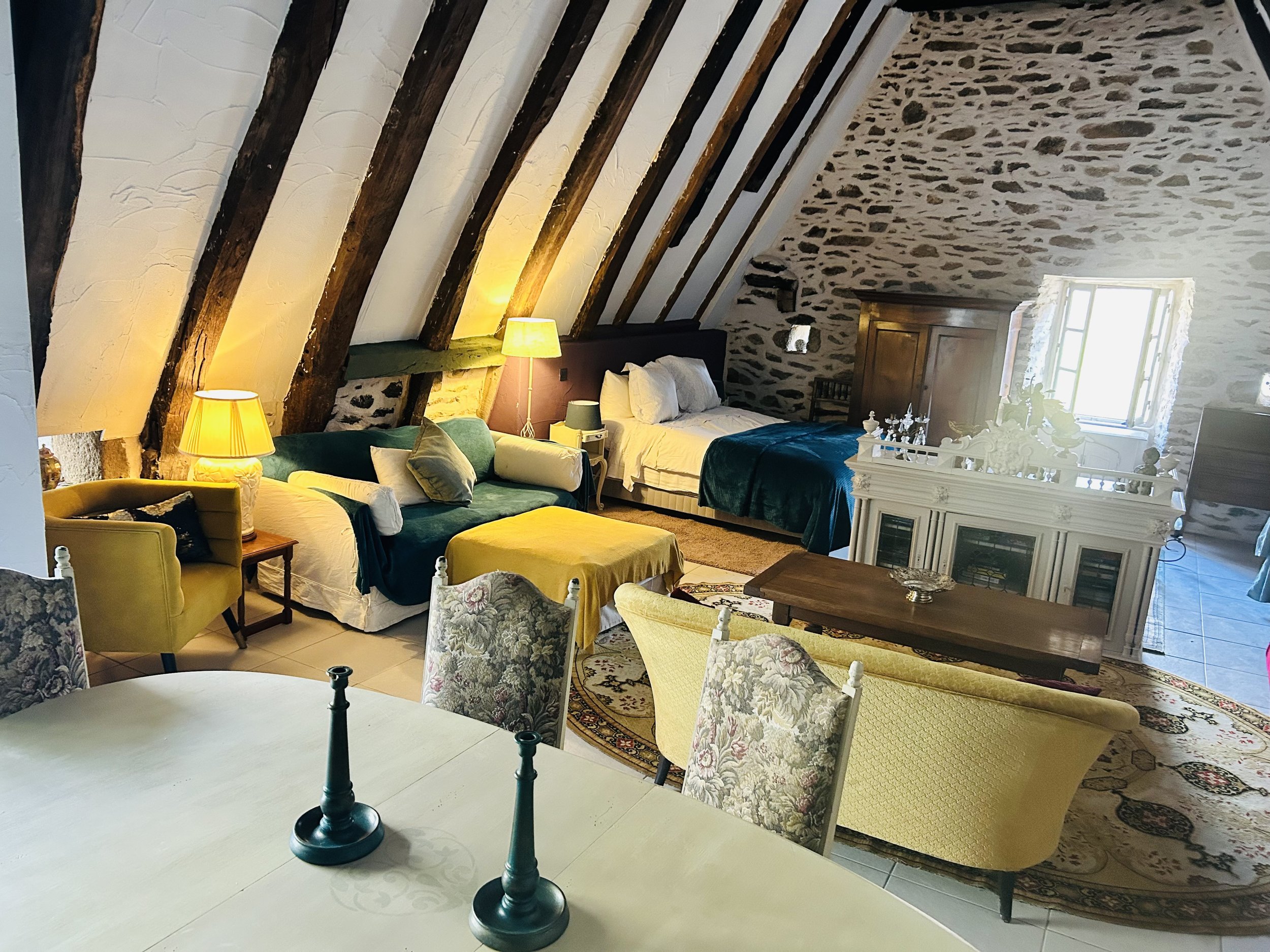 Cozy attic room with slanted wooden beams, stone walls, and rustic decor. Features include a bed, couch with green and yellow cushions, a small dining table, chairs, vintage fireplace, and warm lighting.