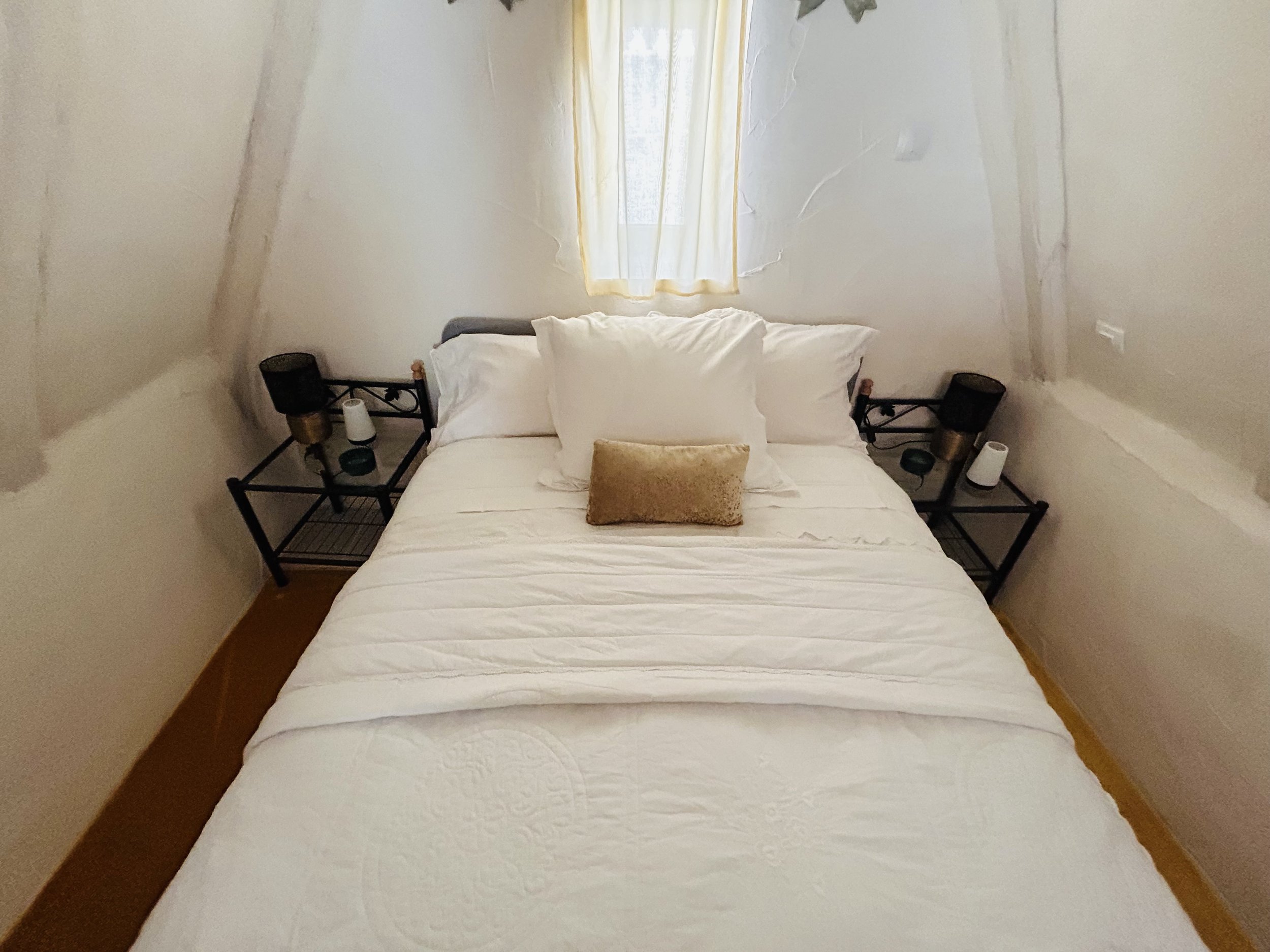 Cozy bedroom with a neatly made bed featuring white bedding, two pillows, and a small decorative cushion. The bed is flanked by nightstands with lamps and accessories. A window with a sheer curtain allows natural light into the room.
