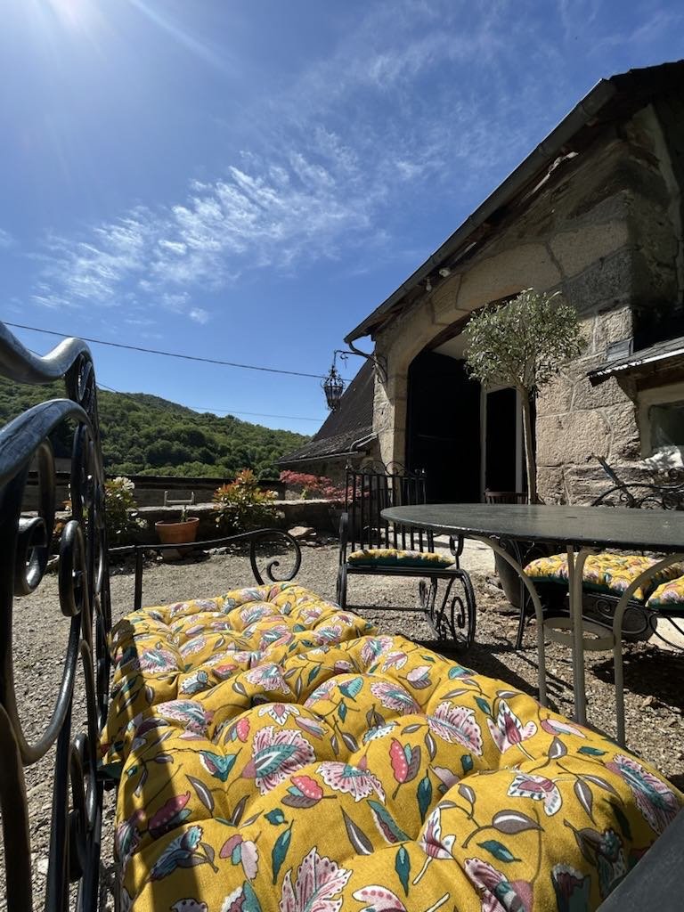 Sunny outdoor patio with floral cushions, iron furniture, rustic stone building, and lush green hillside in the background.