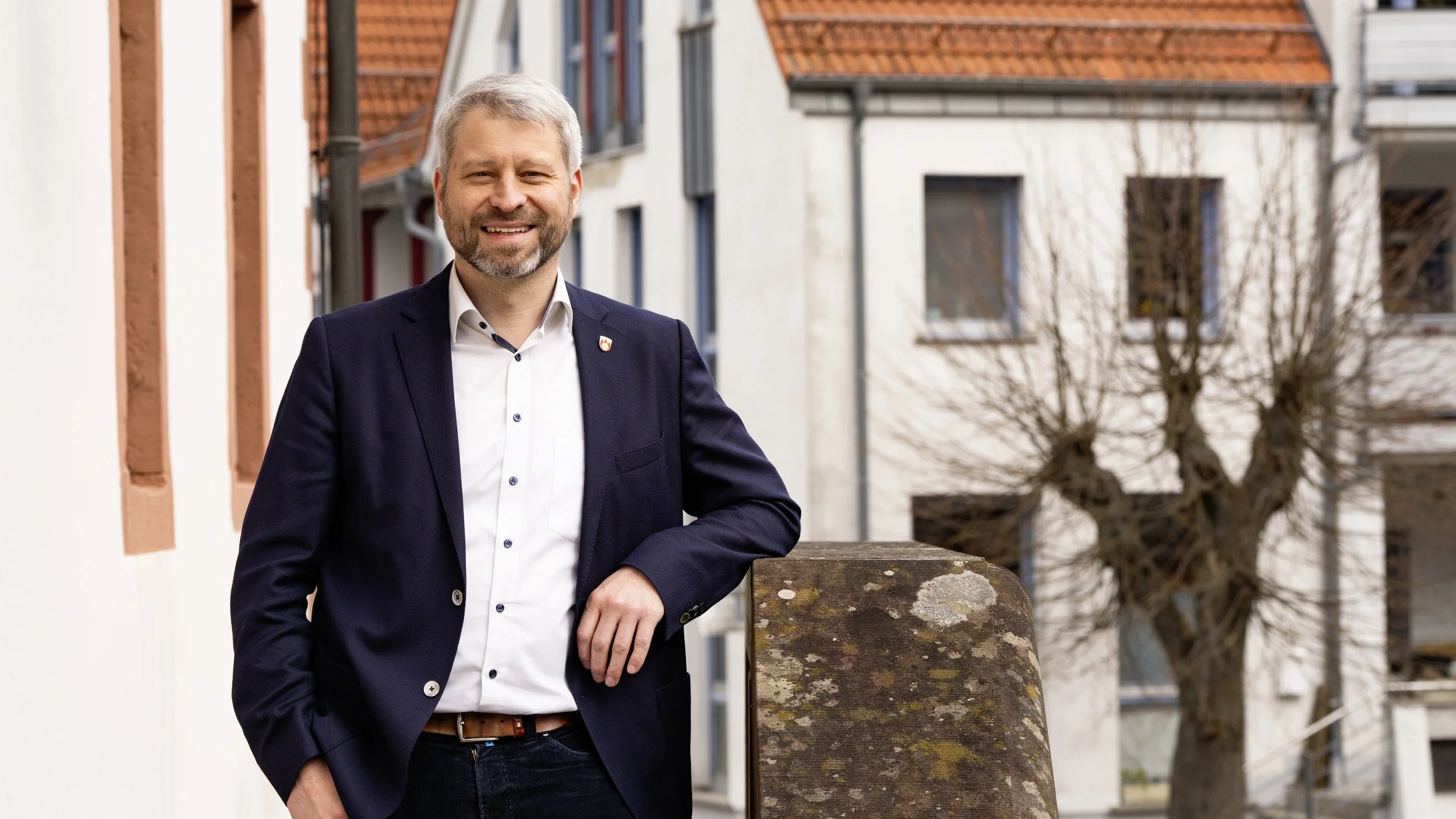 Bürgermeister Peter Kirchner aus Ehrenberg (Rhön). Mann mit grauen Haaren in dunklem Sakko lehnt an einer Steinbalustrade vor einem Gebäude mit roten Dächern und kahlen Bäumen im Hintergrund