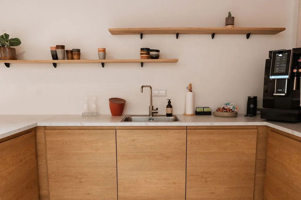 Een moderne keuken met houten kasten, een wit werkblad, open houten planken met potten en planten, een gootsteen, een waterfles, een zeepdispenser, een papieren handdoekhouder, een kom met snacks, en een koffiezetapparaat.