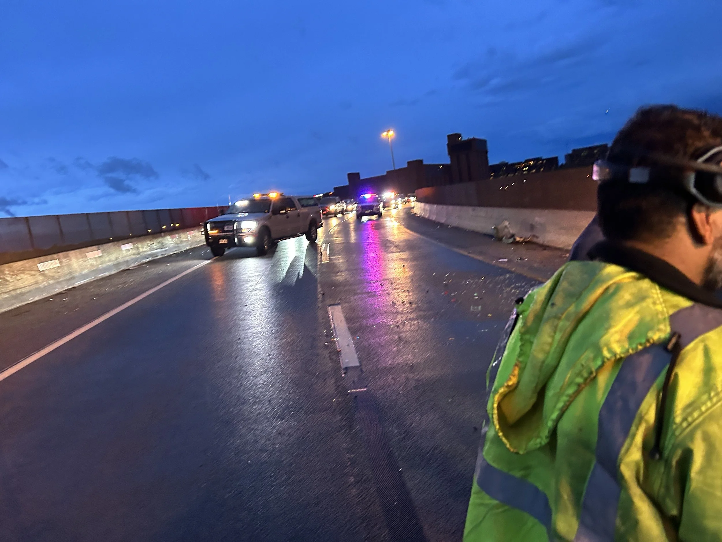 Scene of a wet highway at dusk with traffic stopped, a person wearing a high-visibility jacket in the foreground, and an overcast sky.
