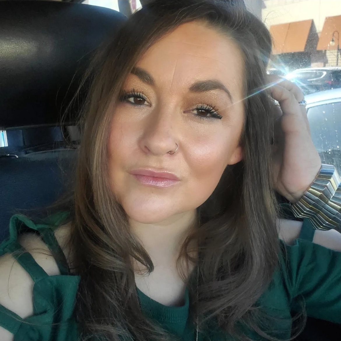 A woman with long brown hair and a nose piercing taking a selfie inside a car, with sunlight reflecting on the window in the background.