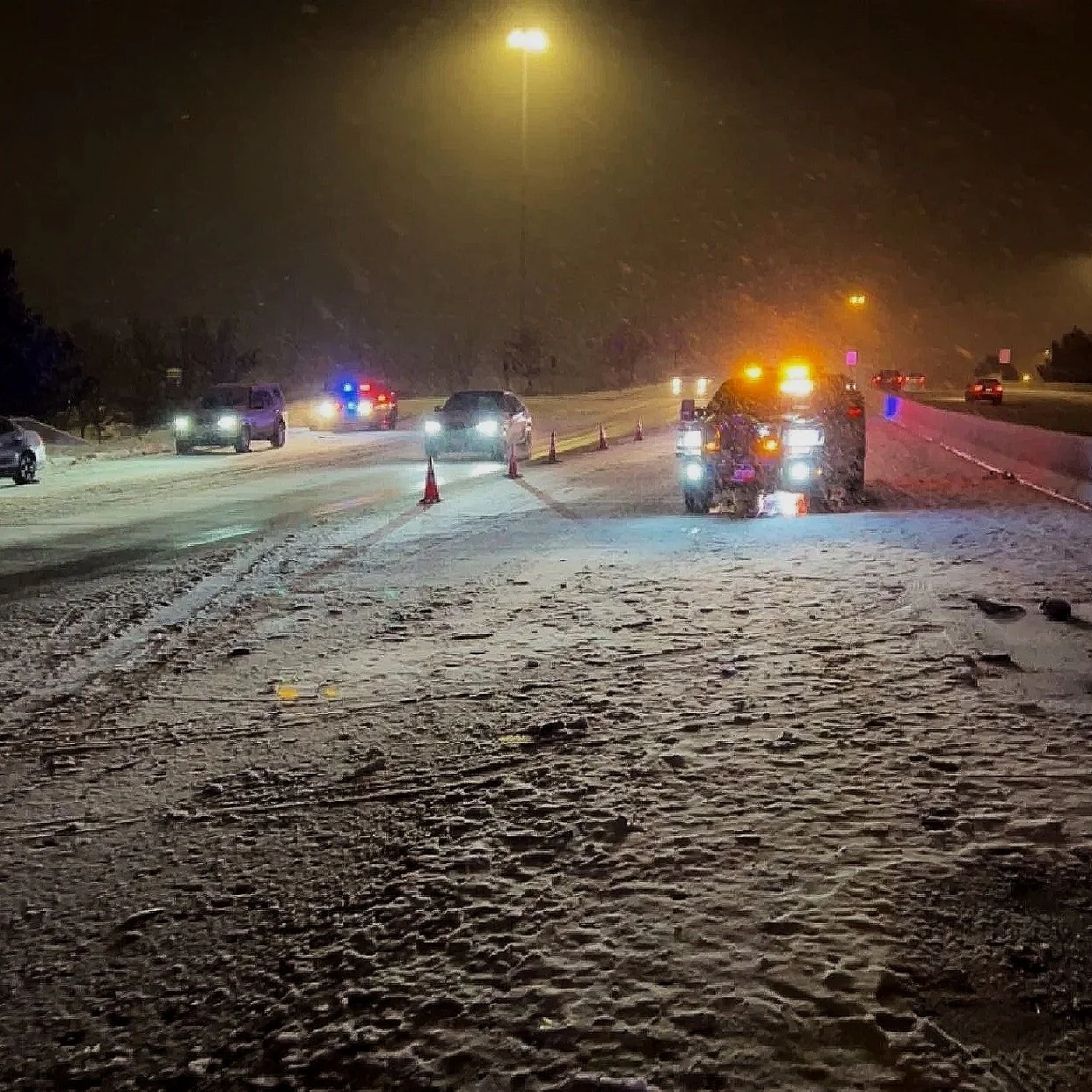 Nighttime scene on a snow-covered road with multiple vehicles, some with flashing lights, and construction cones indicating road work or an accident.